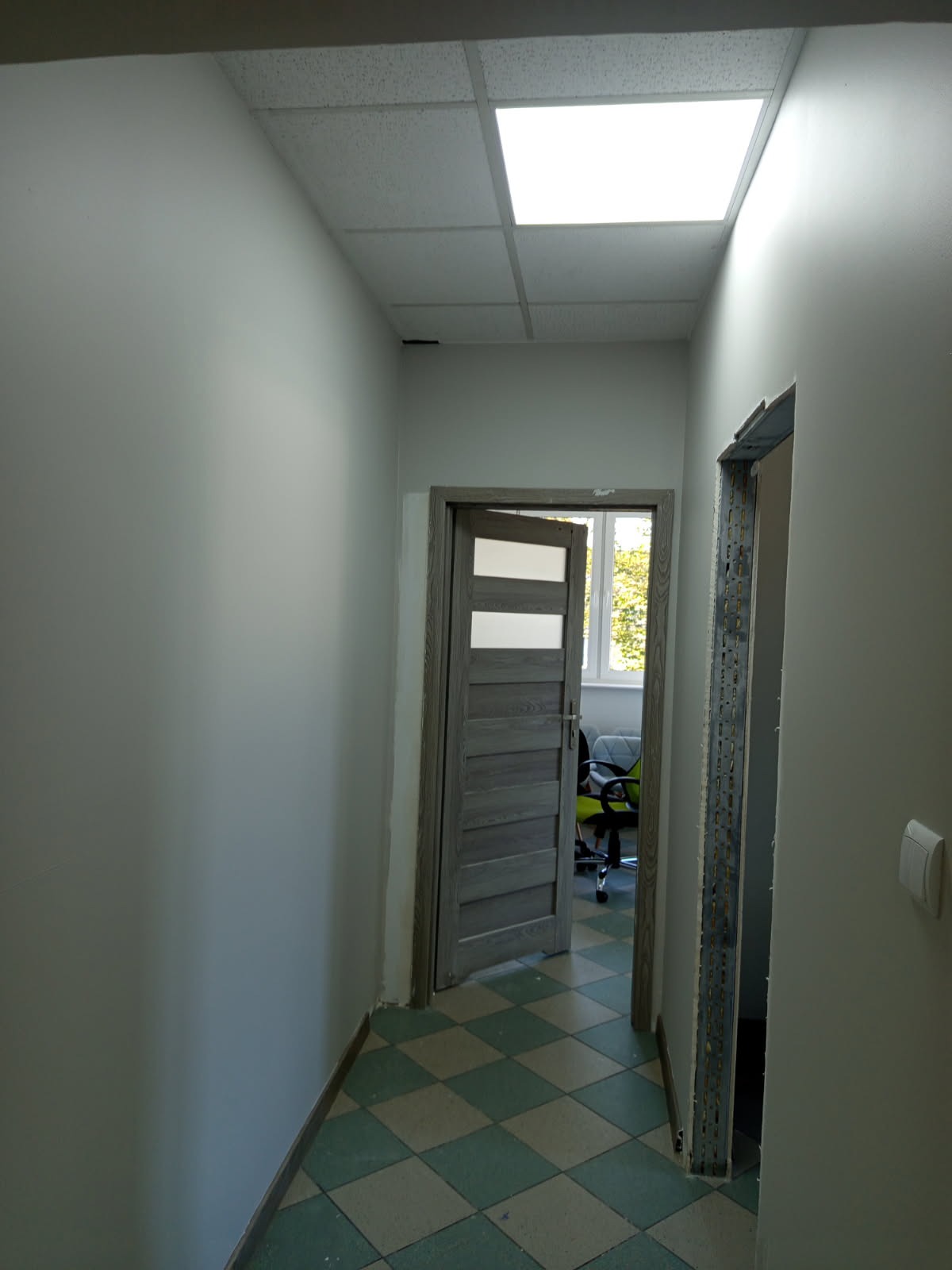 Korytarz po remoncie: szare ściany, podłoga w zielono-białą kratę, dwoje drzwi, jedne otwarte z widokiem na biuro, drugie w trakcie montażu ościeżnicy.