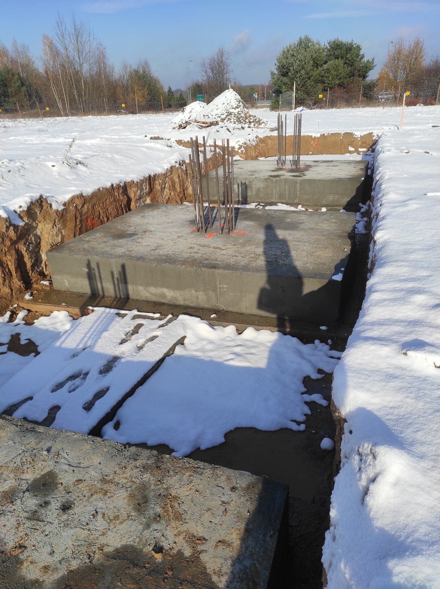 Zimowy krajobraz budowy: wykopane fundamenty z betonu i wystającymi prętami zbrojeniowymi, pokryte śniegiem, w tle drzewa i hałda ziemi.