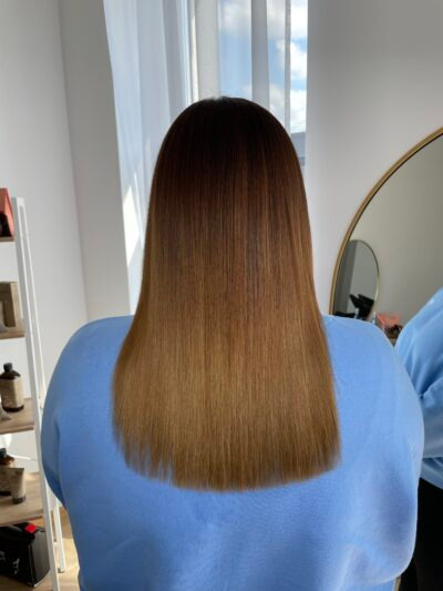 Proste, wygładzone włosy z ombre od ciemnego brązu u nasady do jasnego blondu na końcach, widok z tyłu, w tle biała ściana i okrągłe lustro.