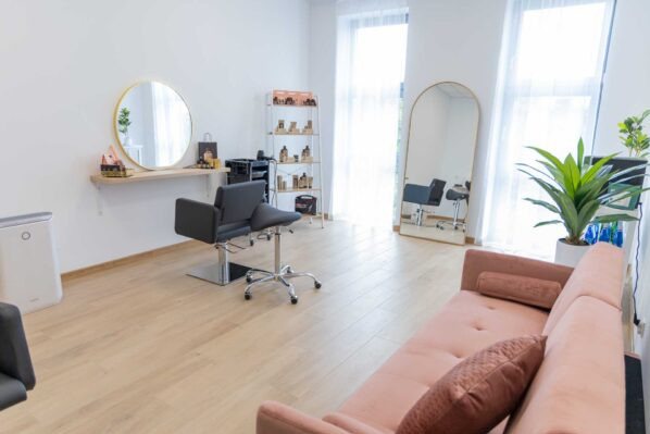 Jasne wnętrze salonu z fotelem fryzjerskim, okrągłym lustrem, regałem z kosmetykami i różową sofą w strefie oczekiwania. Minimalistyczny design z dużymi oknami.