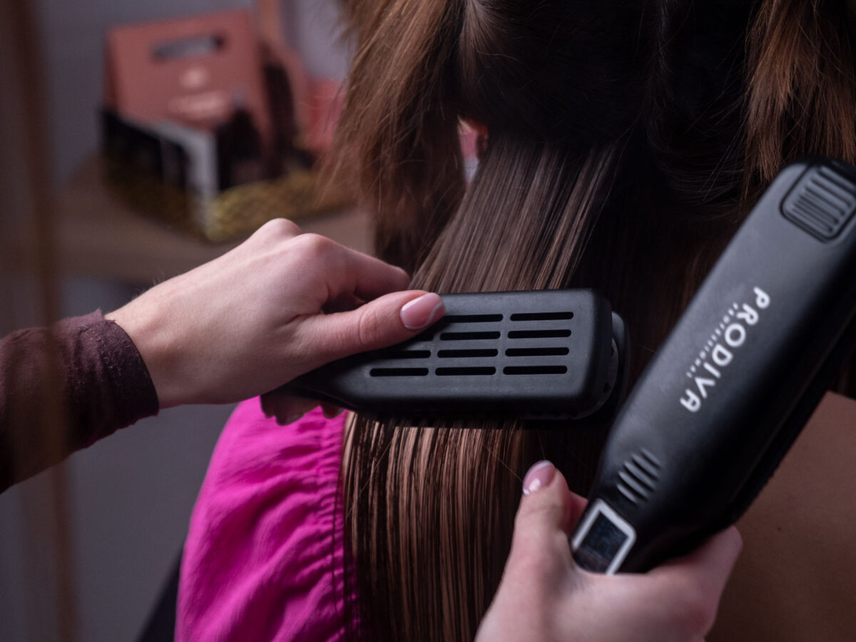 Prostowanie wilgotnych włosów prostownicą marki Prodiva, widoczne dłonie stylistki z delikatnym manicure.