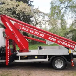 Biały samochód z czerwonym wysięgnikiem koszowym Multitel MT22 z widocznym numerem telefonu, zaparkowany na trawniku w otoczeniu kwitnących drzew.