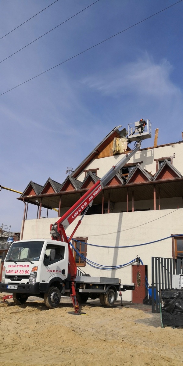 Pracownik w koszu podnośnika na wysięgniku, demontuje elementy dachu budynku z licznymi oknami w szczytach, na pierwszym planie biała ciężarówka z numerem telefonu.