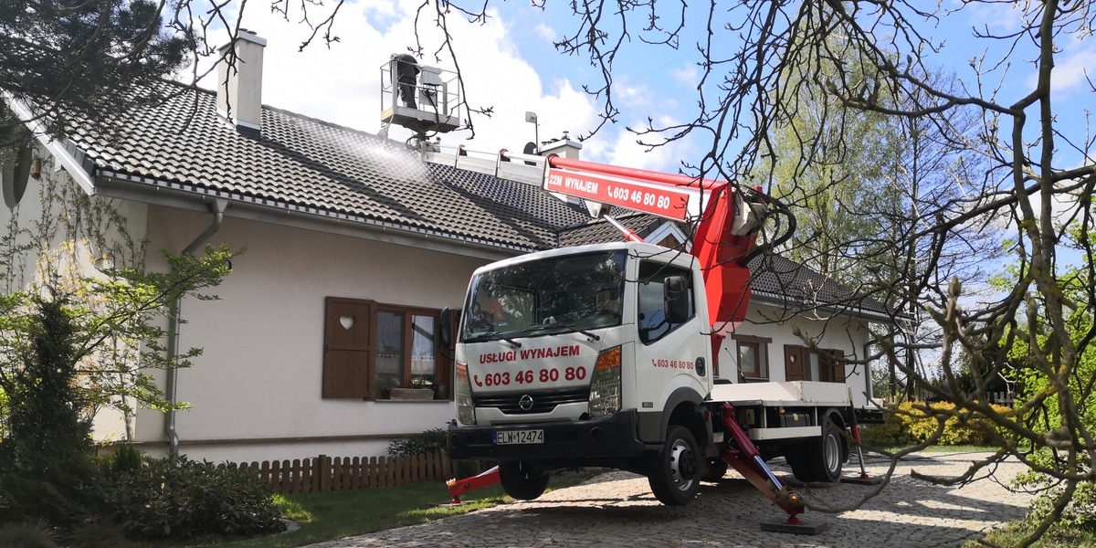 Pracownik na podnośniku koszowym czyści dach domu jednorodzinnego strumieniem wody pod ciśnieniem; widoczny biały samochód z wysięgnikiem i numerem telefonu.