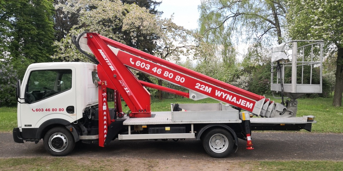 Biały samochód z czerwonym wysięgnikiem koszowym Multitel MT22 z widocznym numerem telefonu, zaparkowany na trawniku w otoczeniu kwitnących drzew.