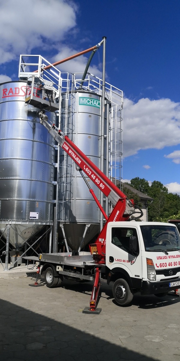 Czerwony podnośnik koszowy na białej ciężarówce ustawiony przed dwoma srebrnymi silosami zbożowymi w słoneczny dzień.