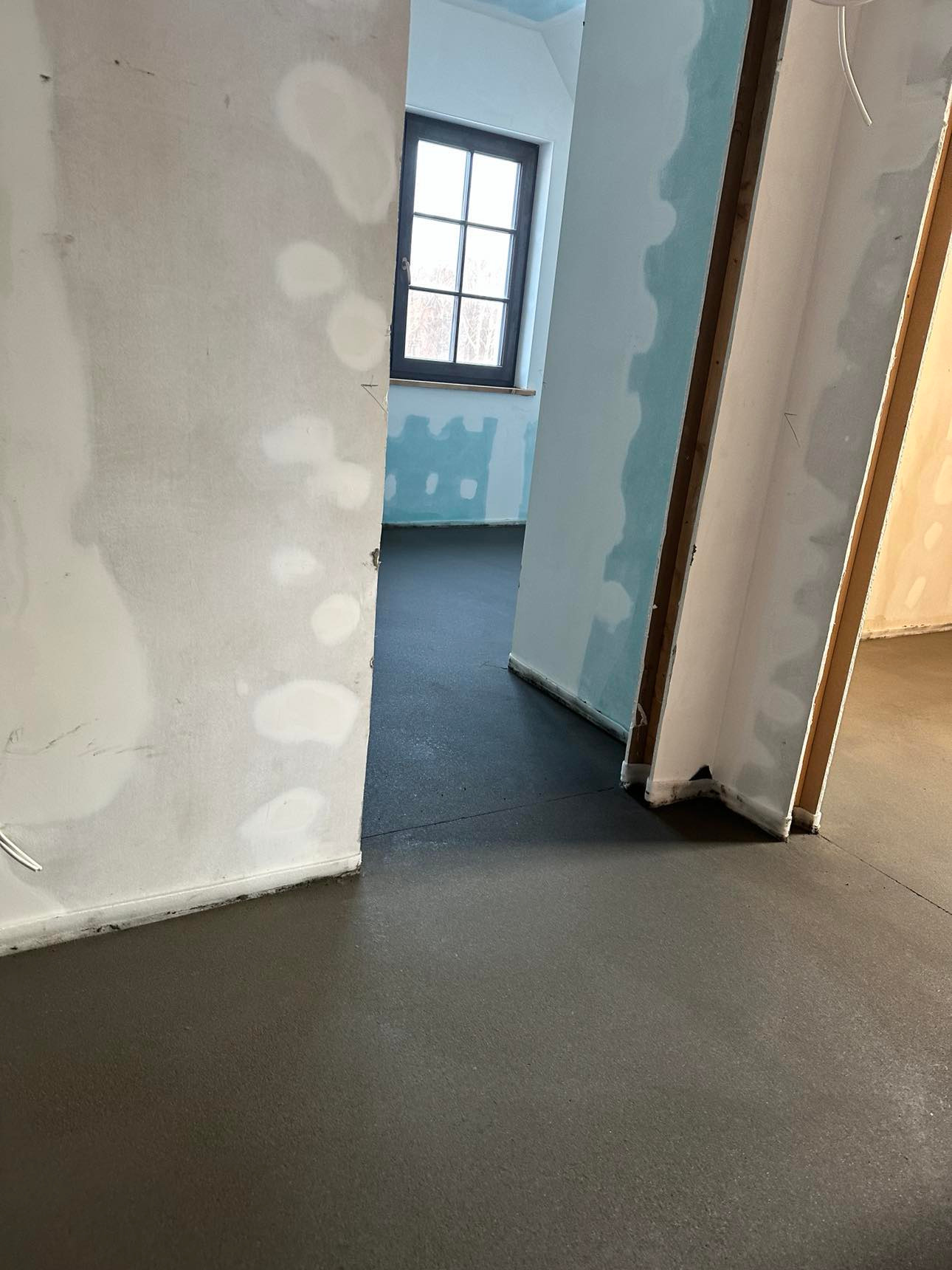 Świeżo wylana, ciemnoszara posadzka w surowym wnętrzu z widocznymi śladami po łataniu ścian, okno z ciemną ramą w tle.