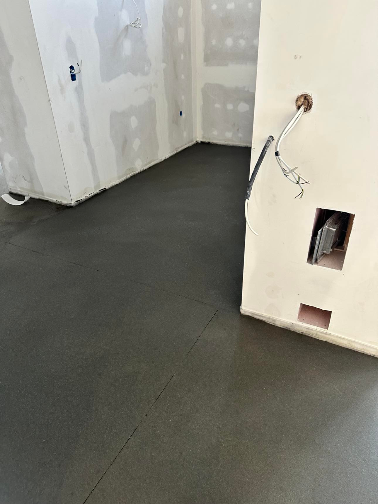 Świeżo wylana, ciemnoszara posadzka w surowym wnętrzu z widocznymi przewodami elektrycznymi i niezabezpieczonymi otworami w ścianie.