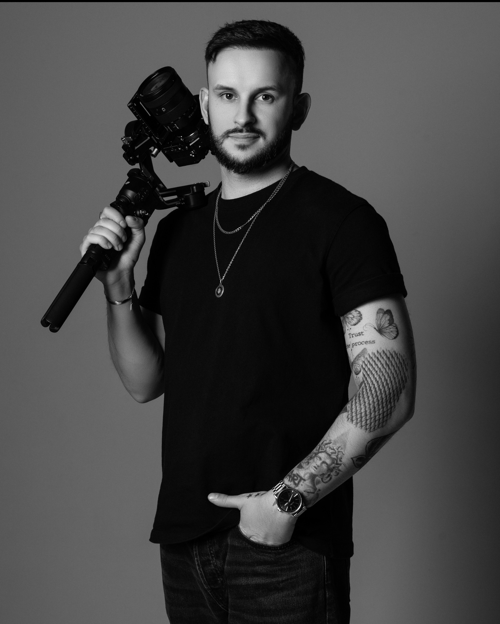 Portret kamerzysty z gimbalem i kamerą, czarno-białe zdjęcie studyjne z widocznymi tatuażami na ramionach i zegarkiem na nadgarstku.