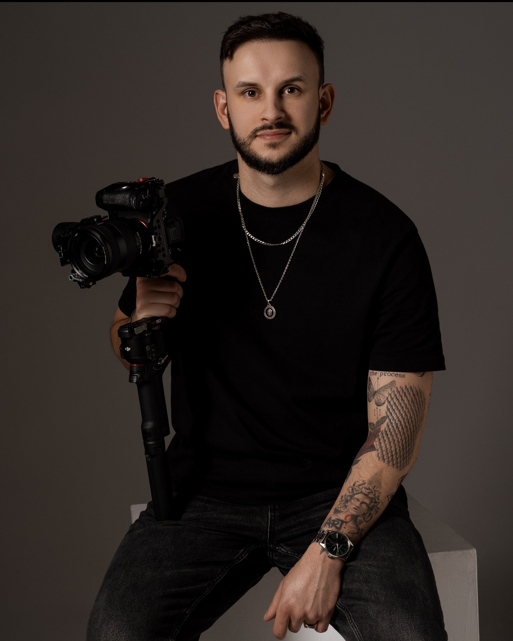 Portret mężczyzny z brodą, w czarnej koszulce i jeansach, trzymającego profesjonalny aparat z gimbalem w dłoni, na szarym tle.