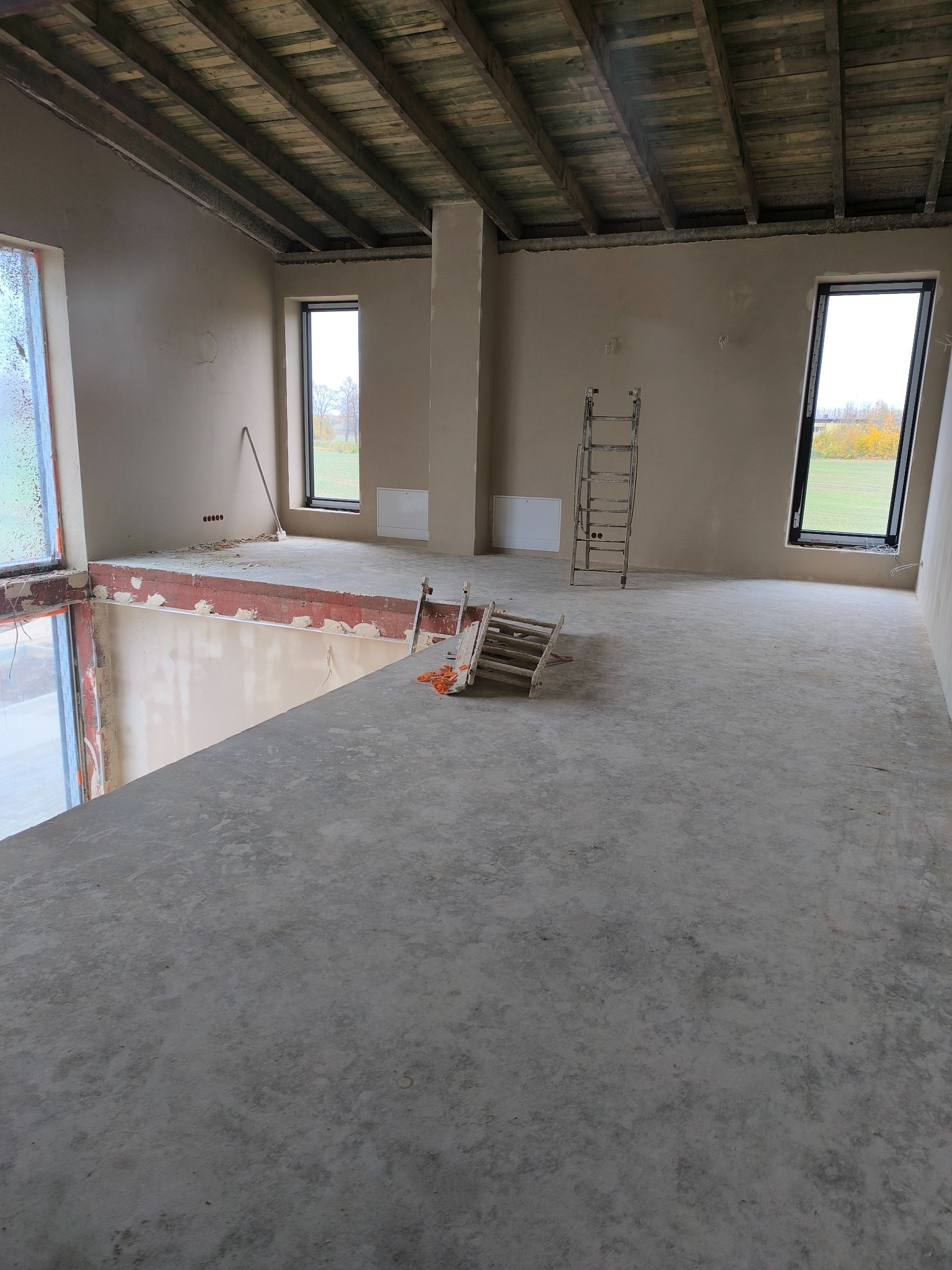 Surowe wnętrze domu w trakcie prac wykończeniowych: ściany po tynkowaniu, surowy beton na podłodze, drabina oparta o ścianę. Widok na zewnątrz przez okna.