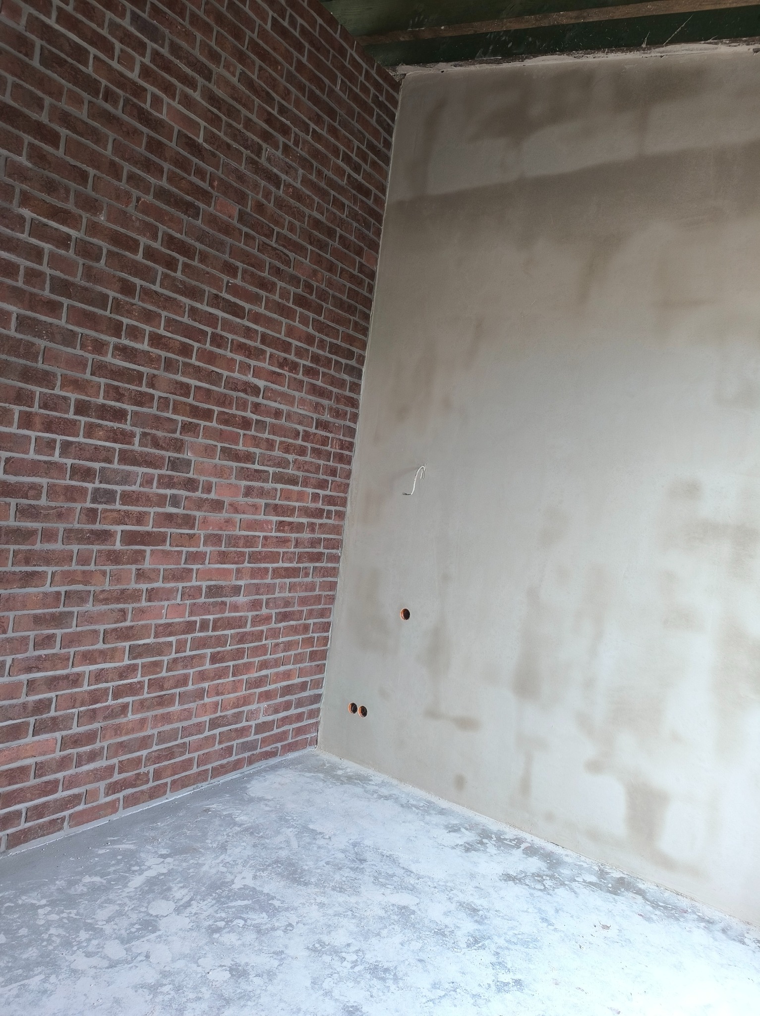 Wnętrze z fragmentem ściany wykończonej czerwoną cegłą i surową, szarą ścianą z widocznymi śladami po szpachlowaniu. Podłoga betonowa, surowa.