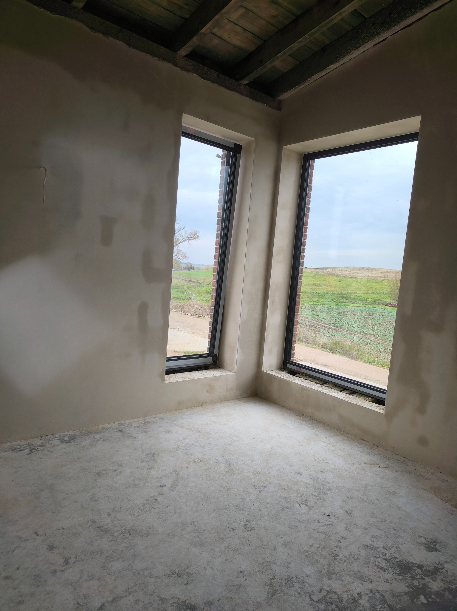 Surowe wnętrze z dwoma oknami z widokiem na pola. Ściany w trakcie wykończenia, sufit z drewnianych belek, betonowa podłoga. Widoczne ślady prac budowlanych.