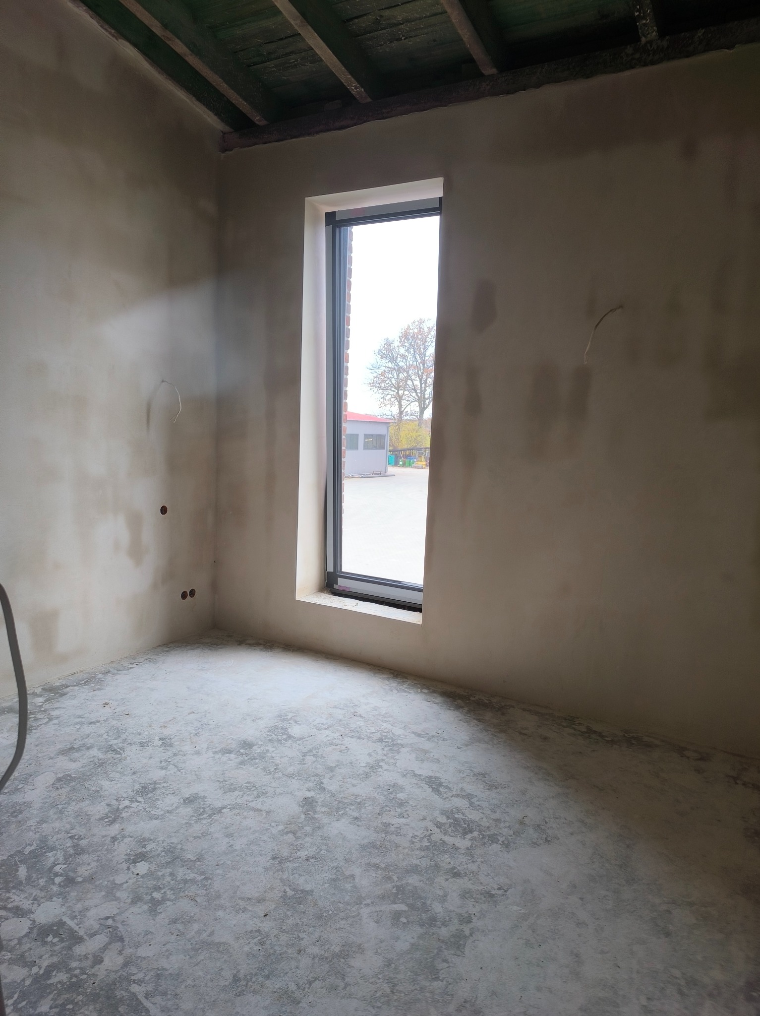 Surowe wnętrze pokoju z oknem, ściany pokryte świeżym tynkiem, widoczne instalacje elektryczne. Podłoga betonowa, sufit z drewnianymi belkami. Światło dzienne wpada przez okno.