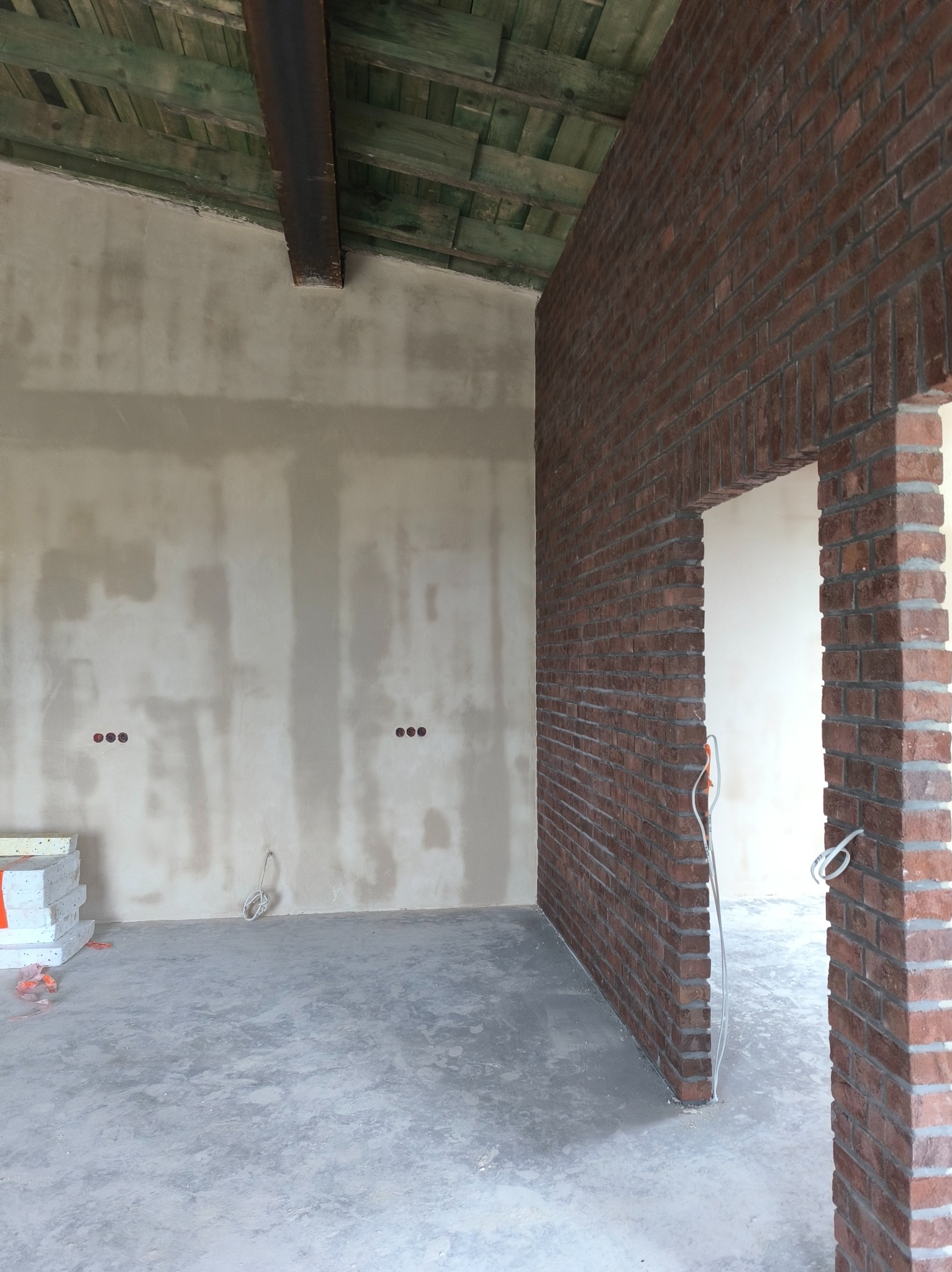 Wnętrze w trakcie prac wykończeniowych: ściana z cegły klinkierowej, surowa ściana z instalacją elektryczną, betonowa podłoga i drewniany strop. Widok na otwór drzwiowy.