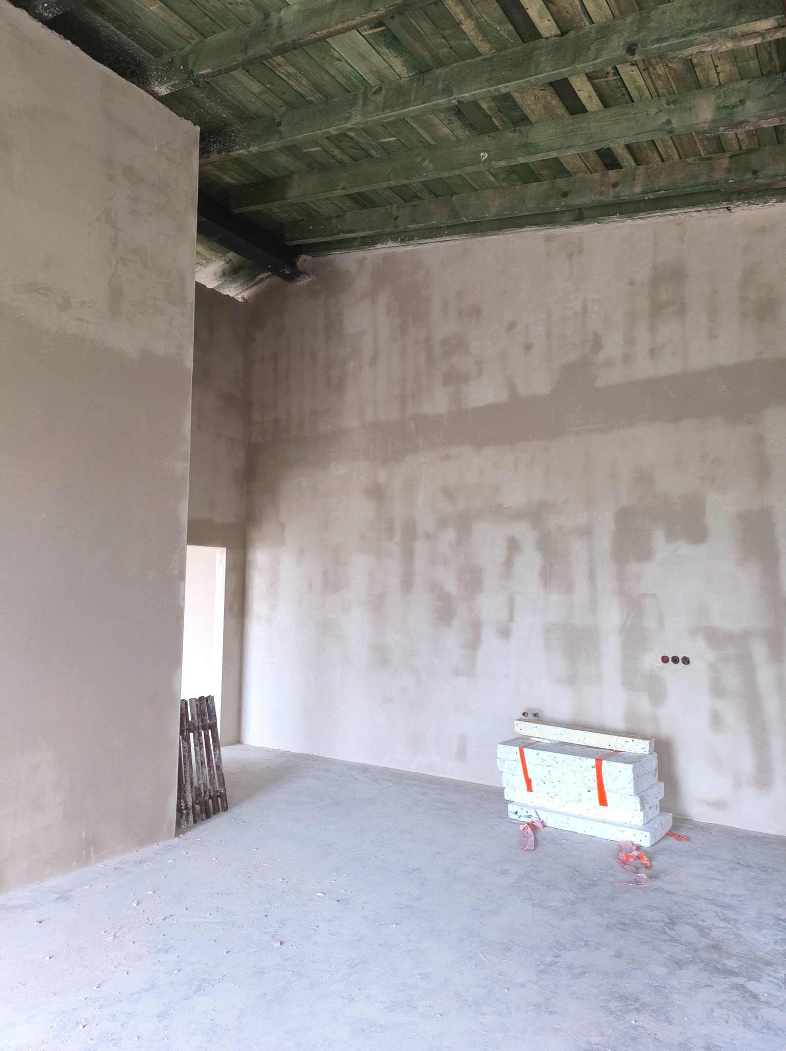 Surowe wnętrze w trakcie prac wykończeniowych: ściany po szpachlowaniu, widoczne instalacje elektryczne, drewniany strop pomalowany na zielono, materiały budowlane na podłodze.