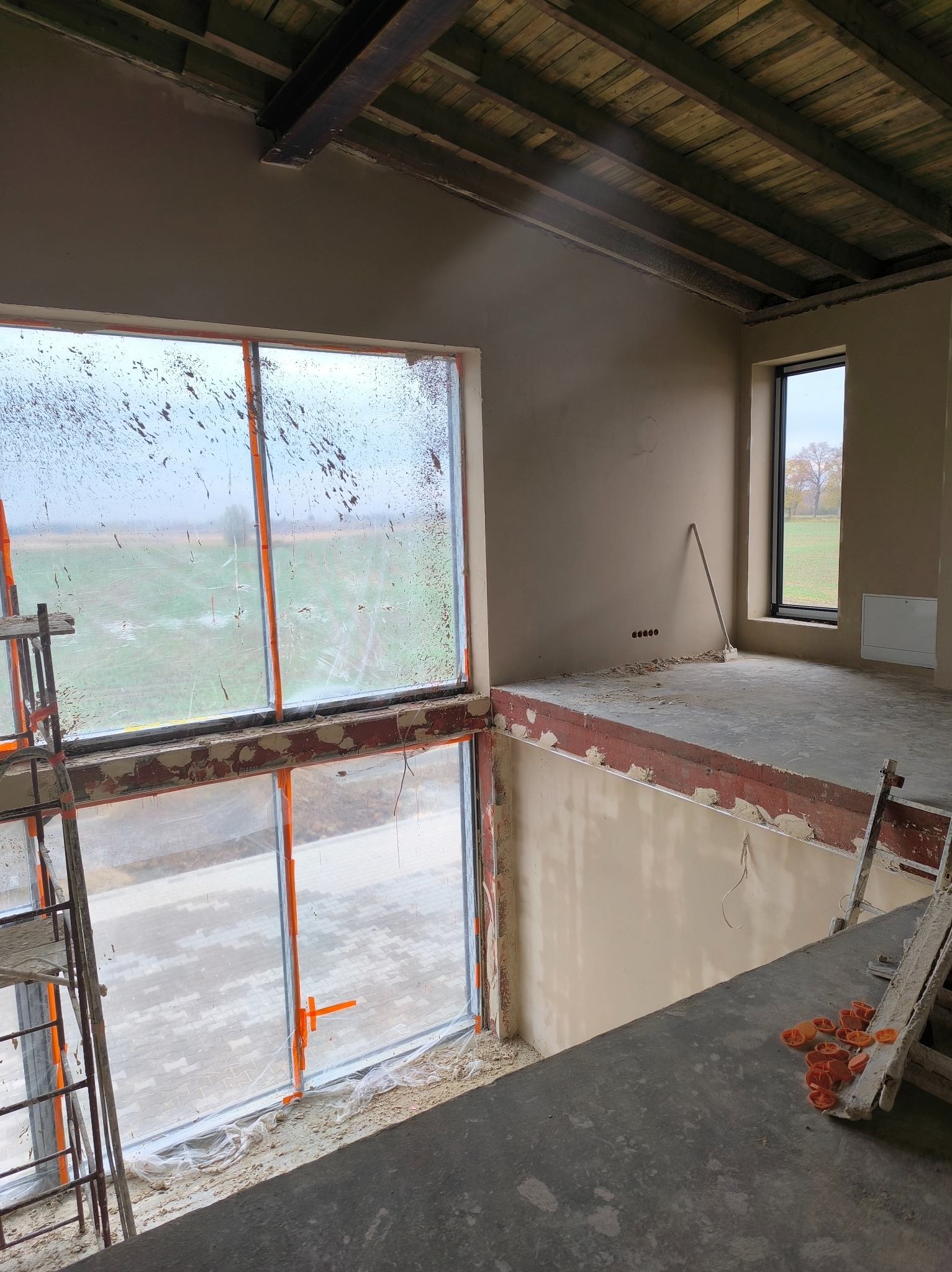 Wnętrze w trakcie tynkowania: ściany pokryte świeżym tynkiem, okna zabezpieczone folią, widoczne rusztowania i narzędzia. Surowy stan wykończenia, widok z góry.