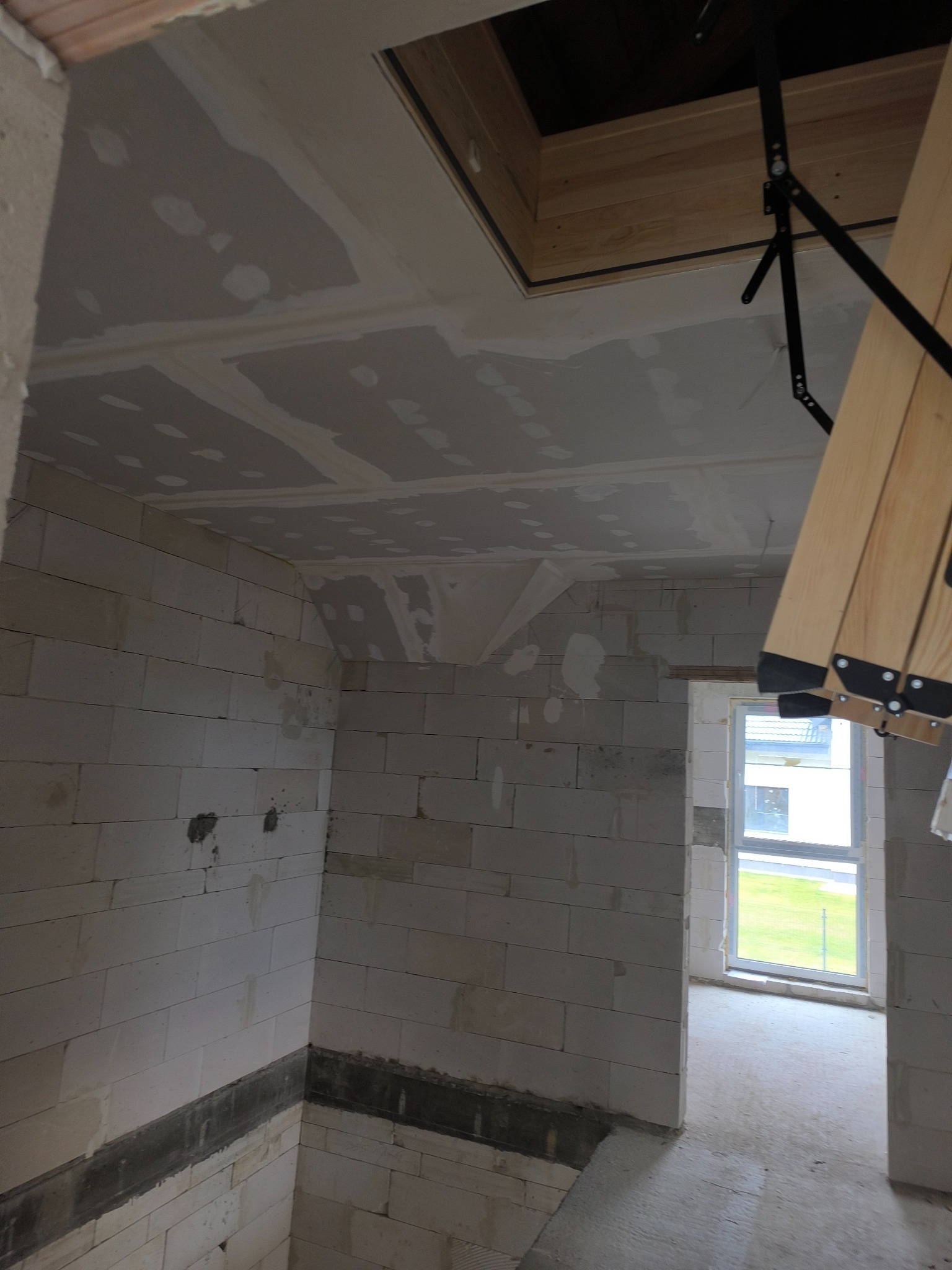 Wnętrze w trakcie szpachlowania: sufit z widocznymi łączeniami płyt gipsowych, ściany z białych bloczków, okno w tle. Widoczne elementy konstrukcji schodów strychowych.