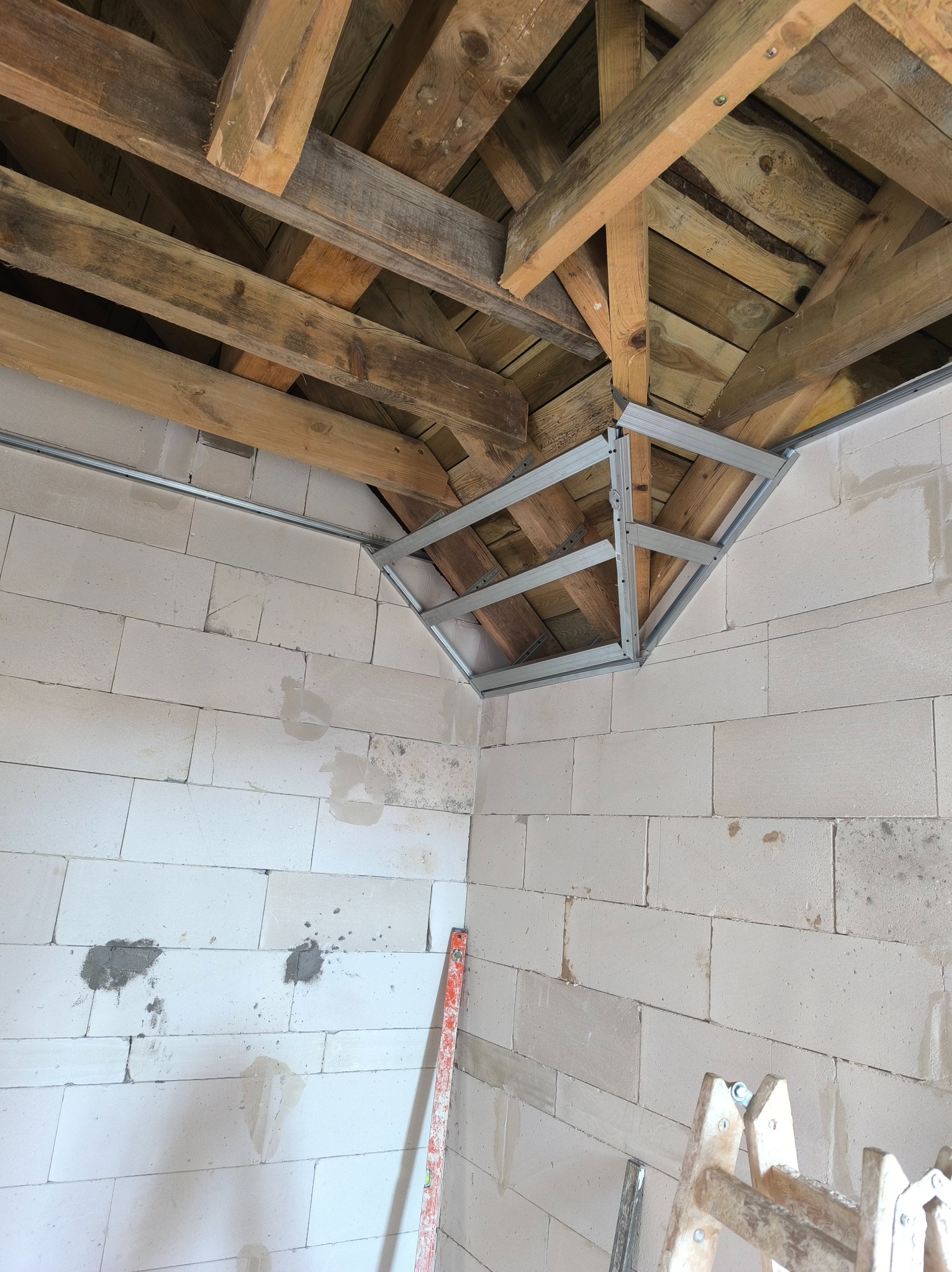 Wnętrze w trakcie prac: sufit z drewnianą konstrukcją i metalowym stelażem, ściany z białych bloczków. Widoczna poziomica i drabina. Przygotowanie do wykończenia.