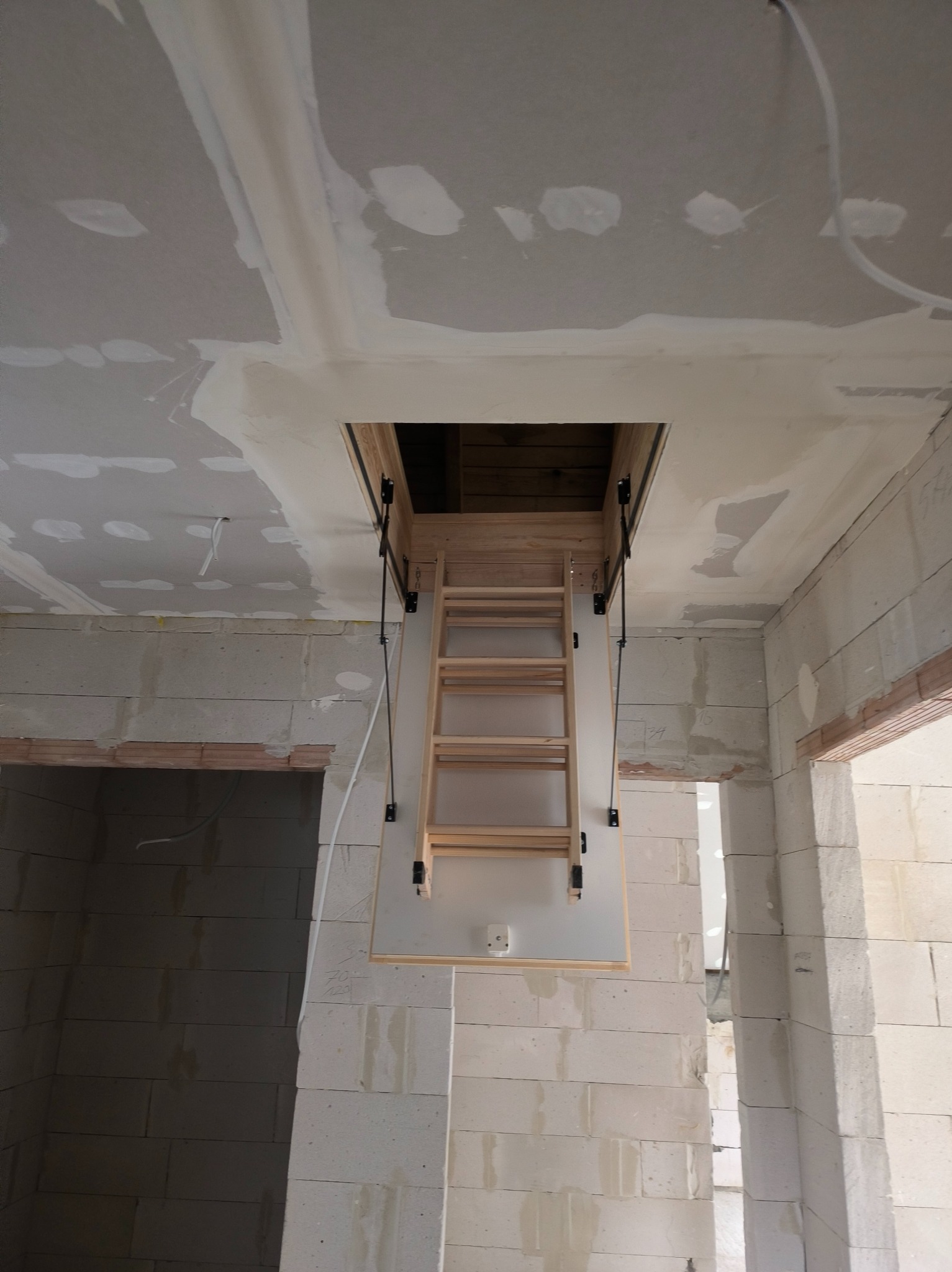 Schody strychowe zamontowane w suficie z widocznymi śladami szpachlowania w Olsztynie. Ściany z białych bloczków, surowy stan wykończenia.