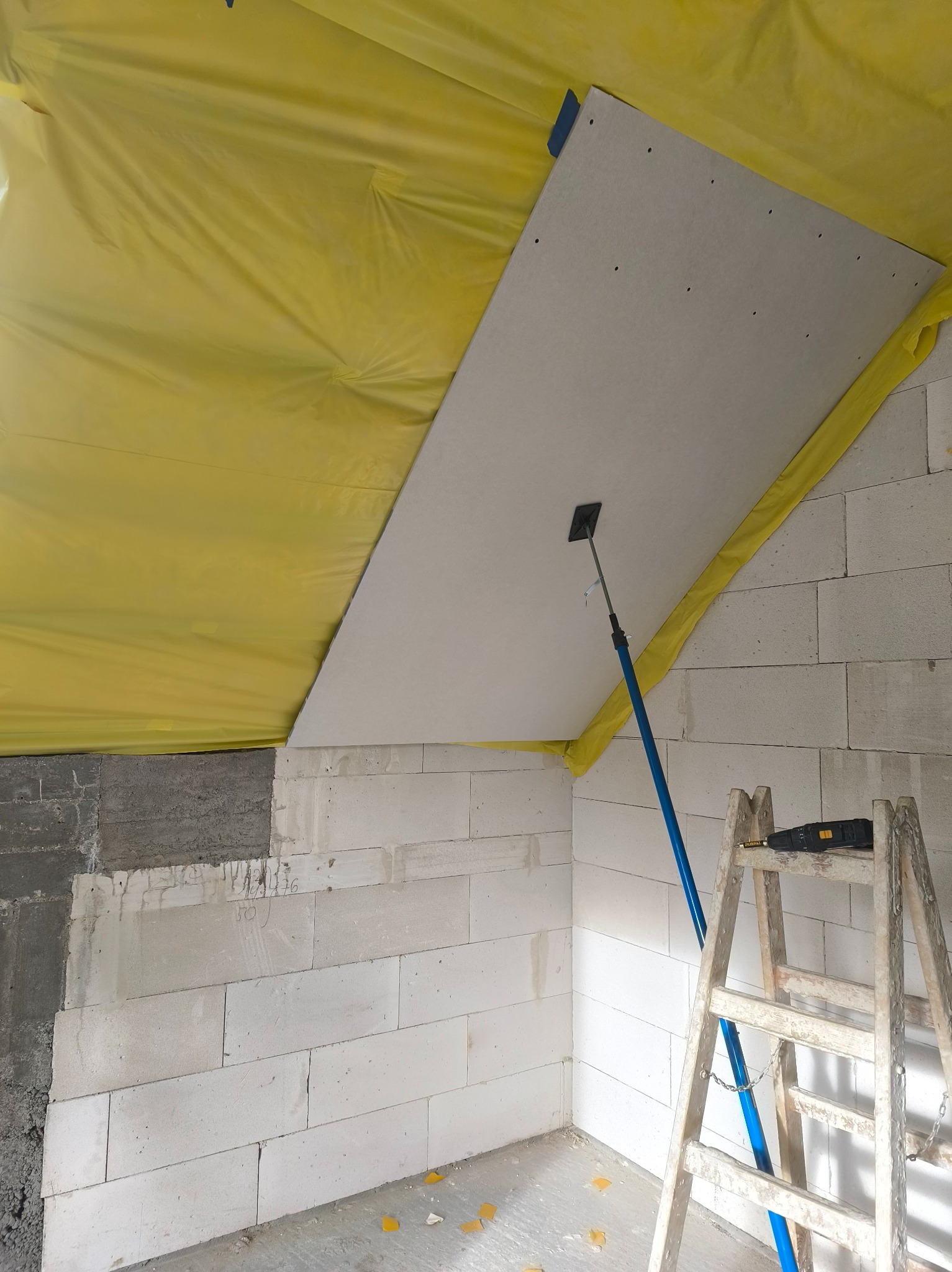 Montaż płyty gipsowej na suficie poddasza, widoczna drabina i podpórka teleskopowa. Żółta folia zabezpiecza przed zabrudzeniem. Ściany z białych bloczków.