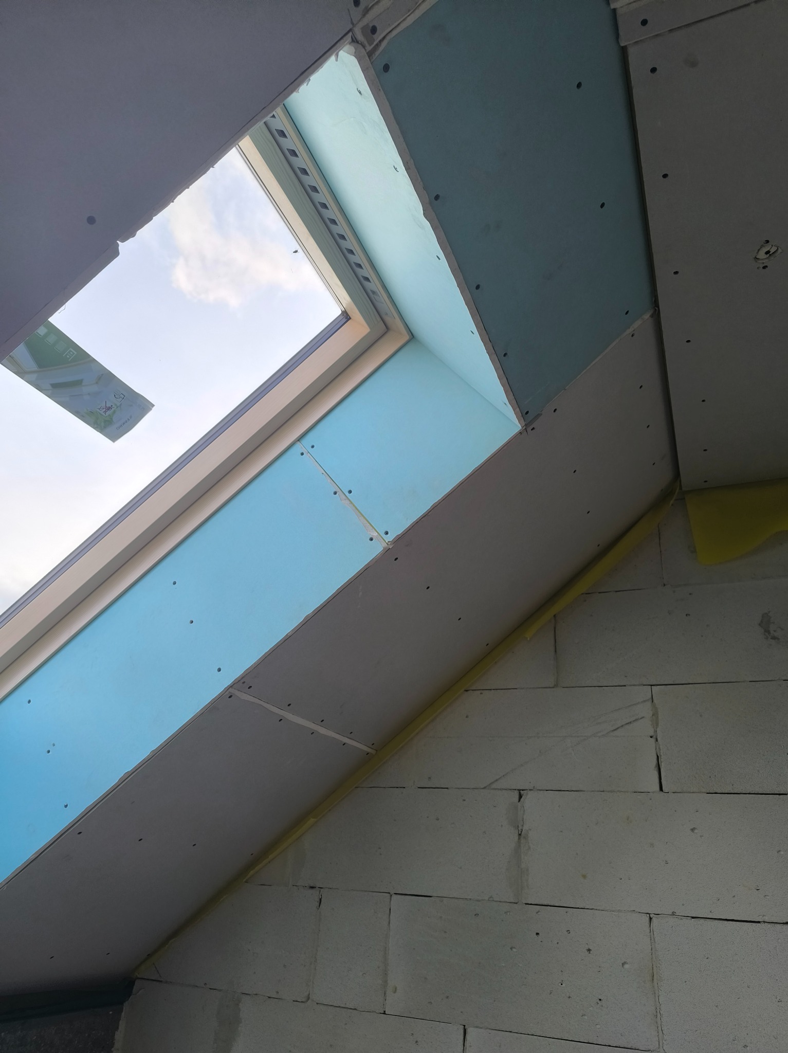 Montaż płyt gipsowo-kartonowych wokół okna dachowego, widoczne łączenia i śruby, ściana z białych bloczków. Etap prac wykończeniowych.