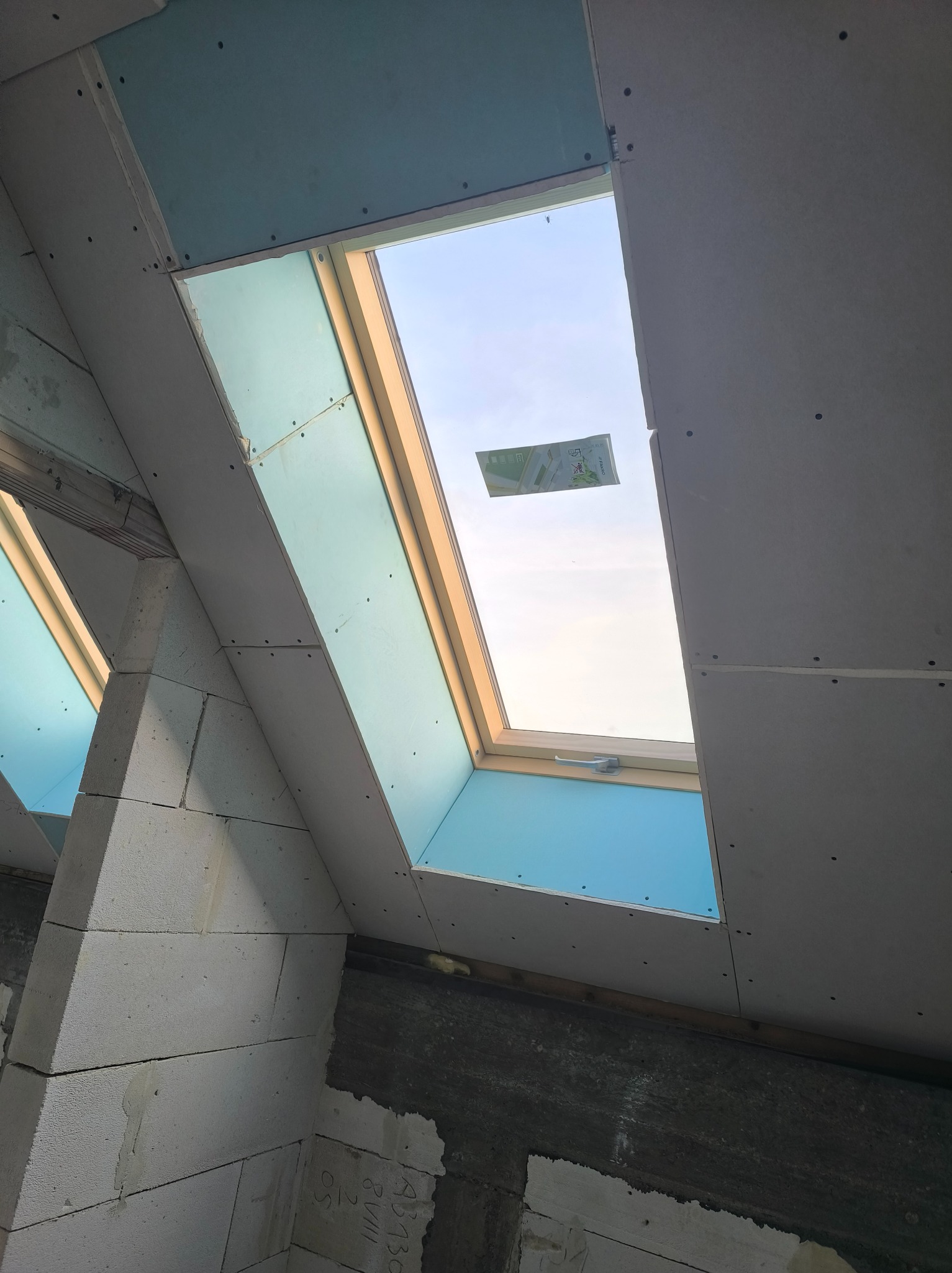 Montaż okna dachowego w skośnej ścianie poddasza, widoczne płyty gipsowo-kartonowe i bloczki z betonu komórkowego. Etap prac wykończeniowych.