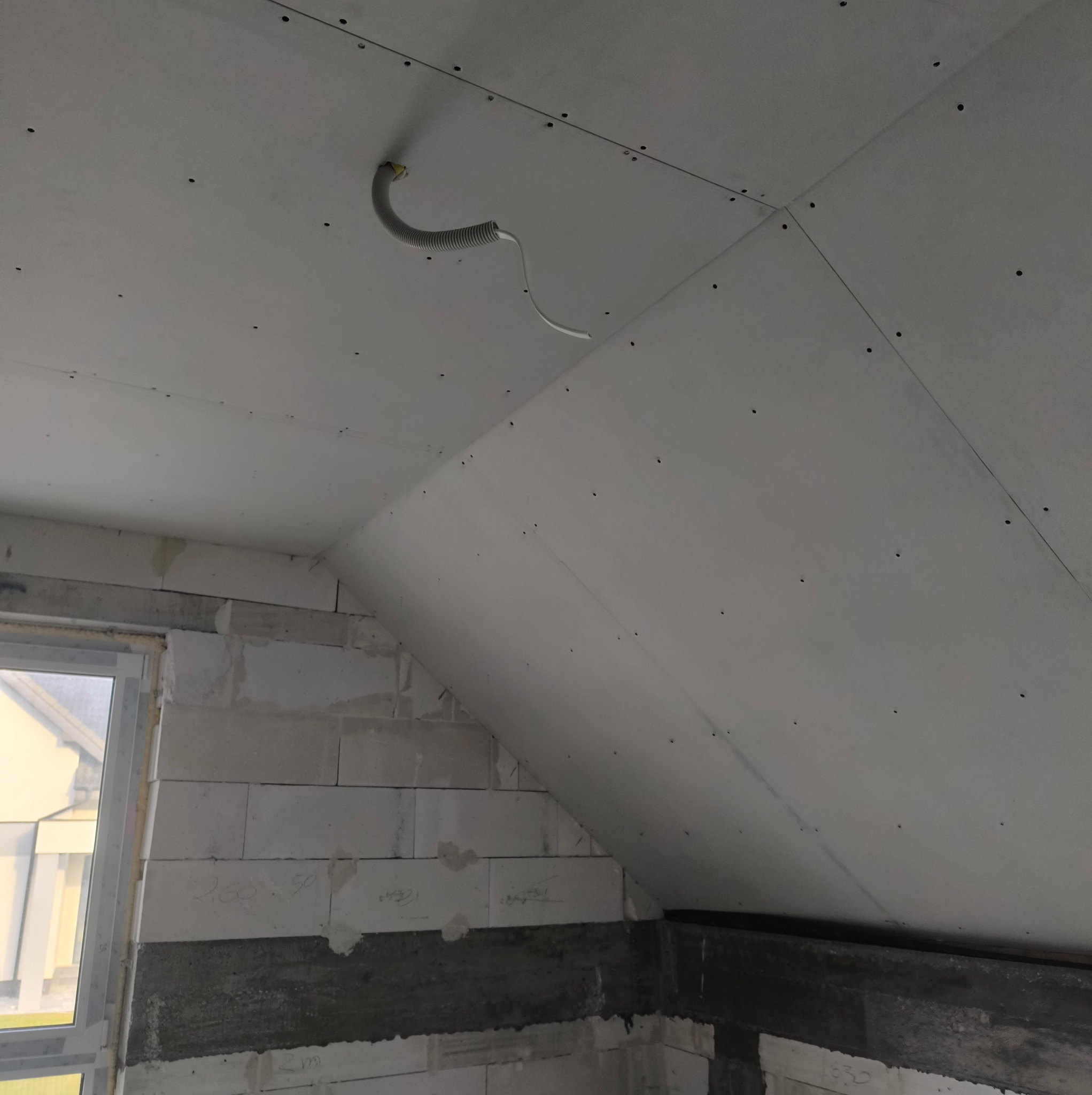Skośny sufit poddasza z płyt gipsowo-kartonowych, widoczne łączenia i wkręty, fragment instalacji elektrycznej w peszlu, ściana z betonu komórkowego przy oknie.