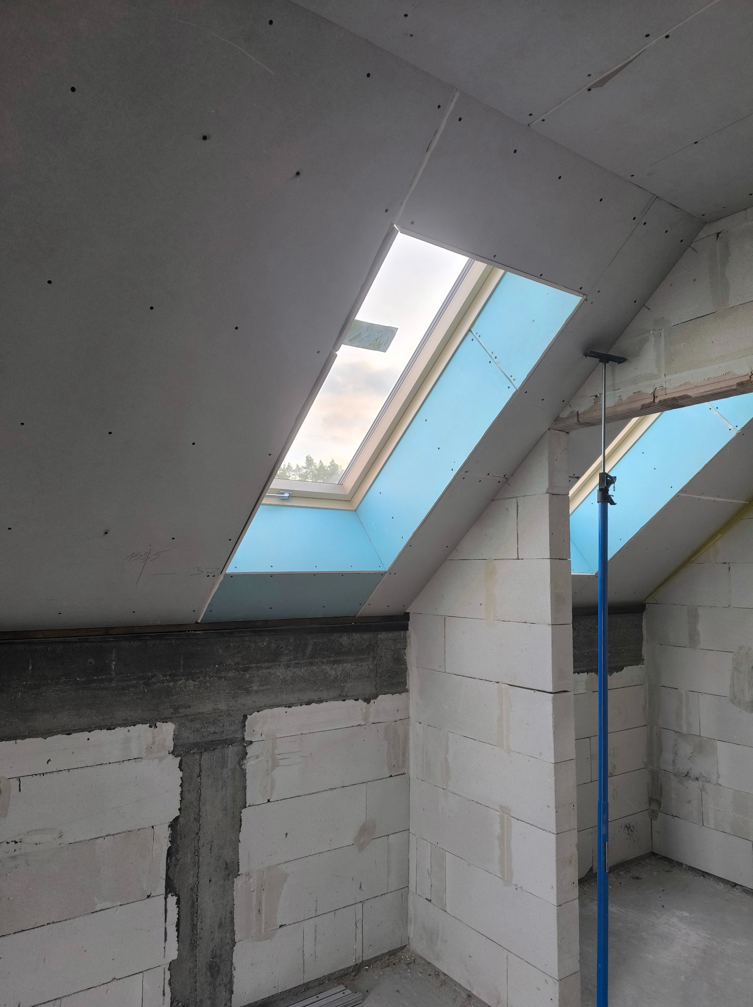Poddasze w trakcie wykańczania: widoczne okna dachowe, ściany z białych bloczków i płyty gipsowo-kartonowe na suficie, podparte niebieską podporą.