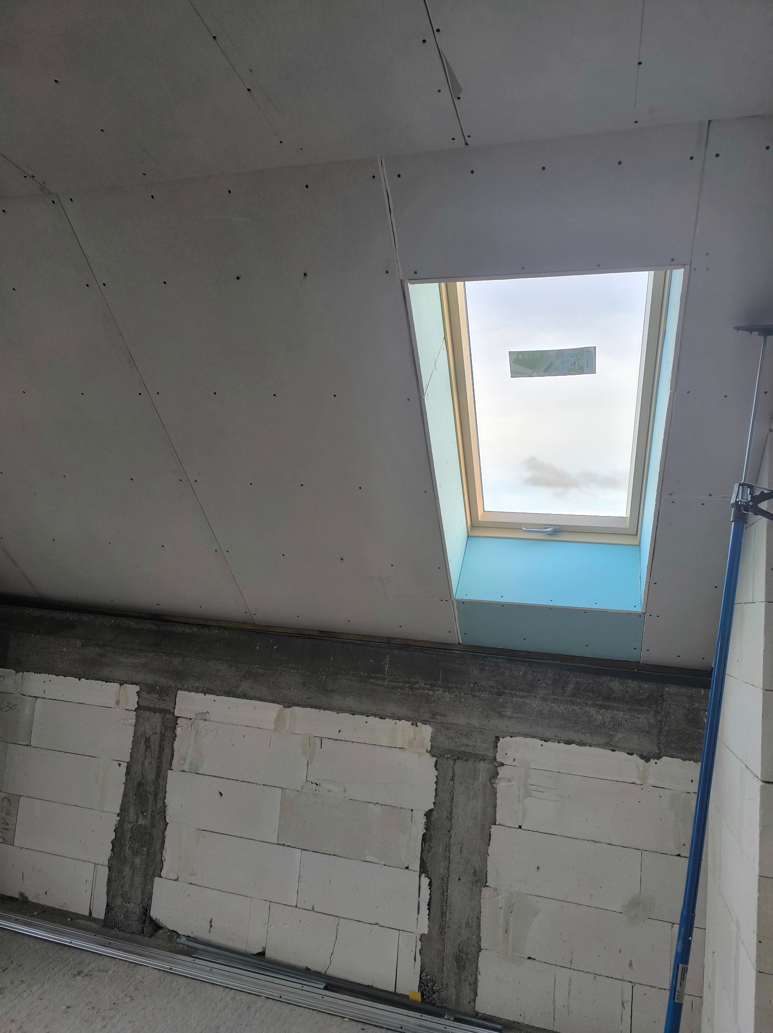 Montaż płyt gipsowo-kartonowych na poddaszu z oknem dachowym. Widoczne łączenia płyt i fragment ściany z bloczków. Prace wykończeniowe w trakcie realizacji.