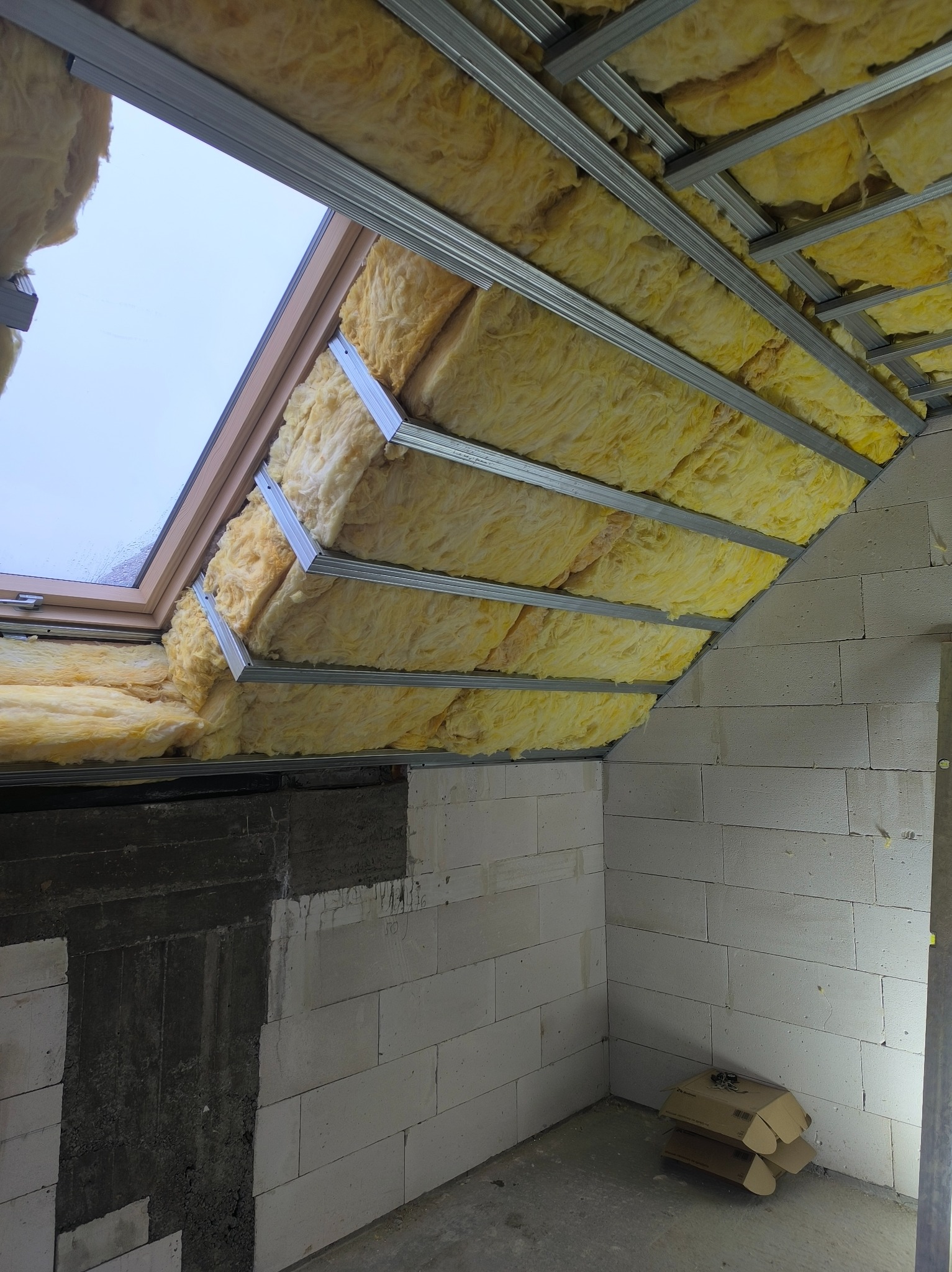 Ocieplenie poddasza wełną mineralną, widoczne metalowe profile i okno dachowe. Ściany z białych bloczków, karton na podłodze. Ujęcie z dołu, surowe wykończenie.