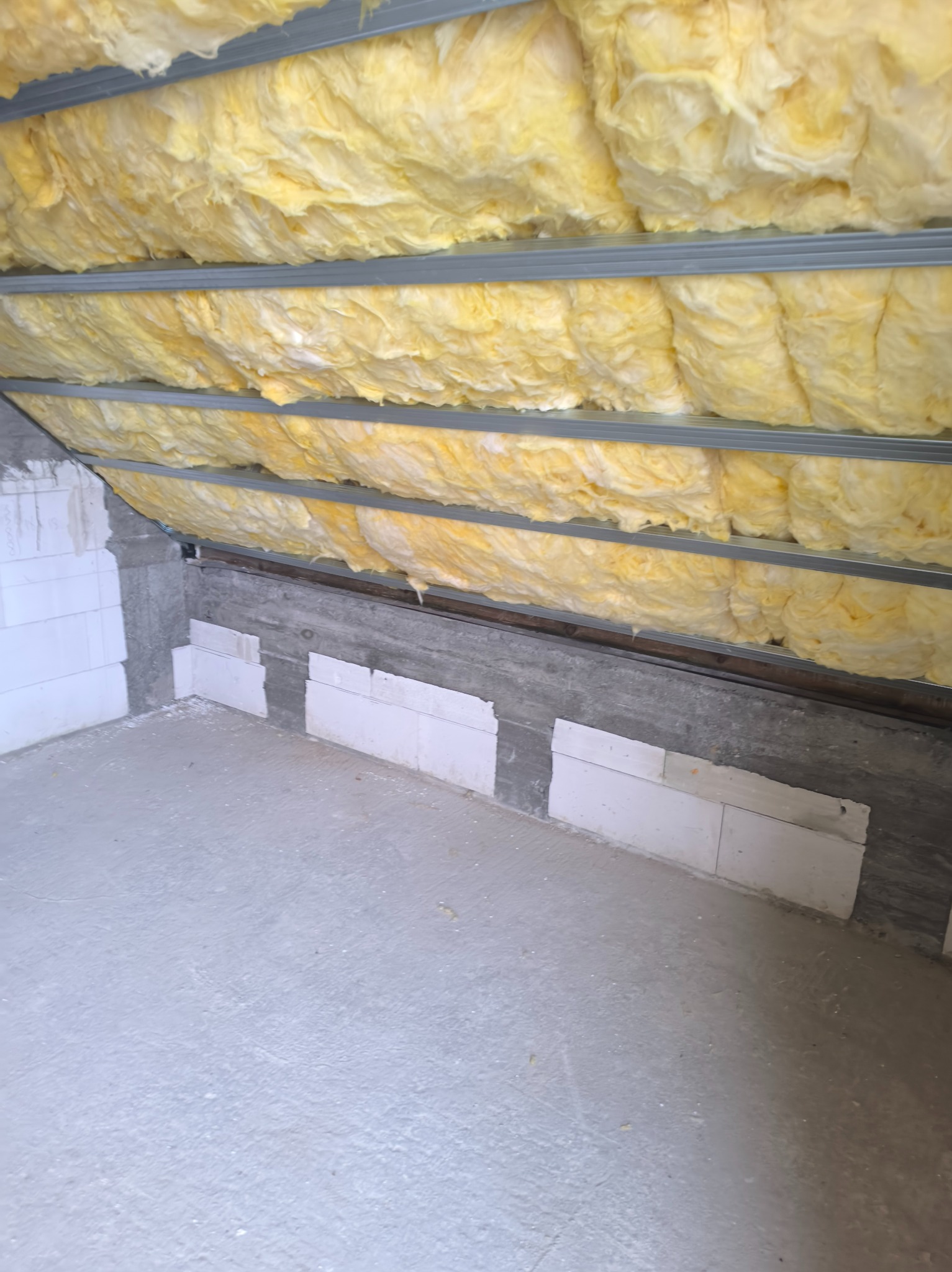 Ocieplenie poddasza wełną mineralną, widoczne metalowe profile i fragment ściany z bloczków. Prace wykończeniowe w surowym wnętrzu.