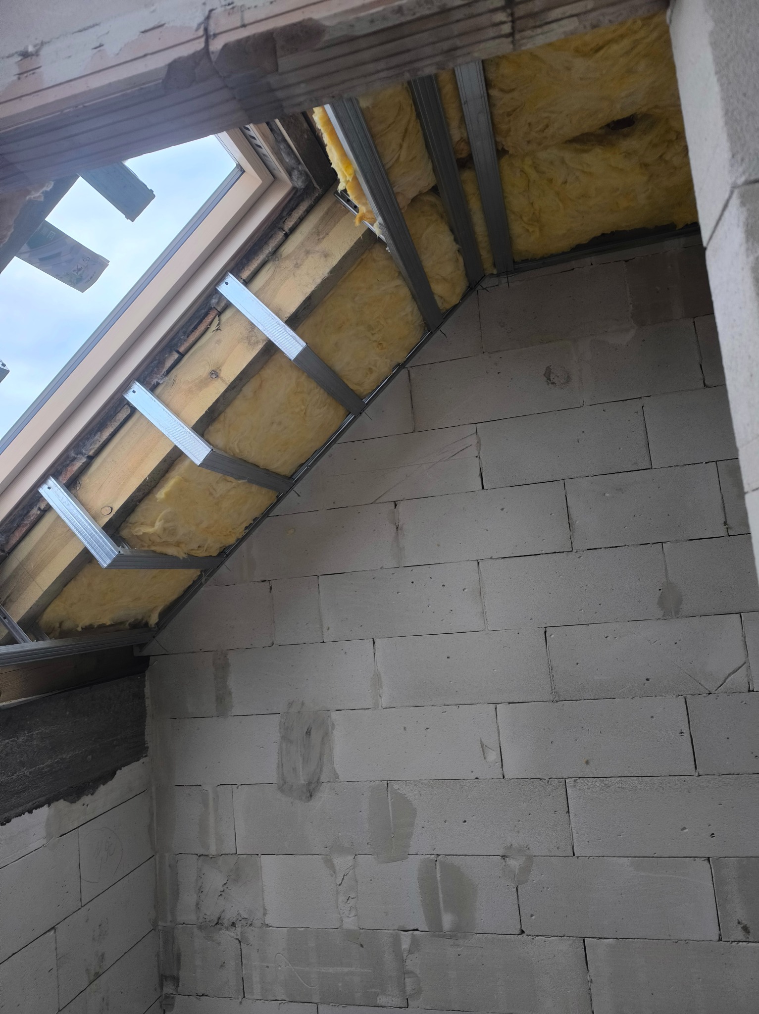 Ocieplenie poddasza wełną mineralną, widoczne metalowe profile i fragment okna dachowego. Ściany z białych bloczków.