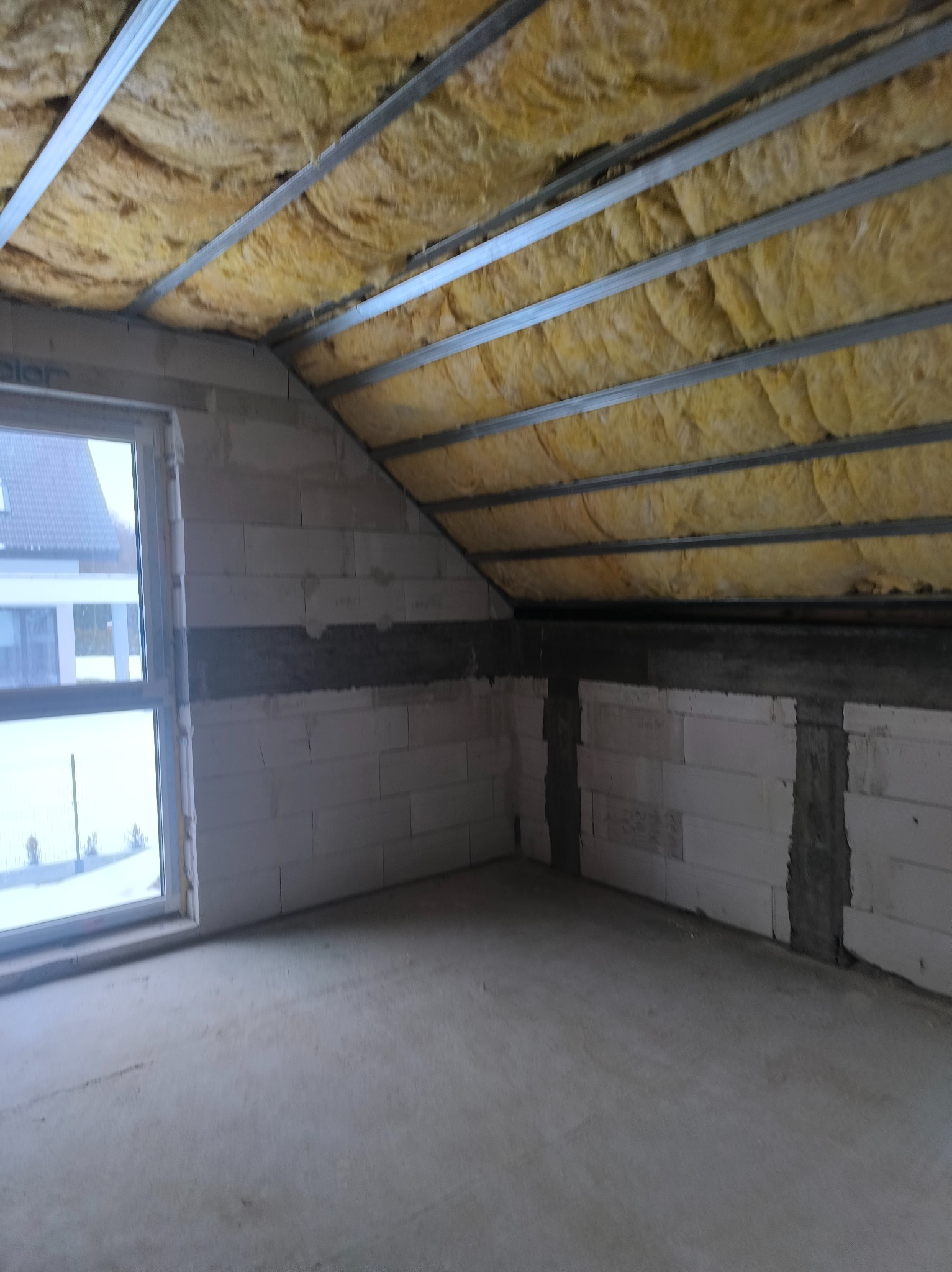 Poddasze w trakcie adaptacji: widoczne ocieplenie wełną mineralną pod metalowym stelażem sufitu, ściany z betonu komórkowego, okno. Surowy stan wykończenia.