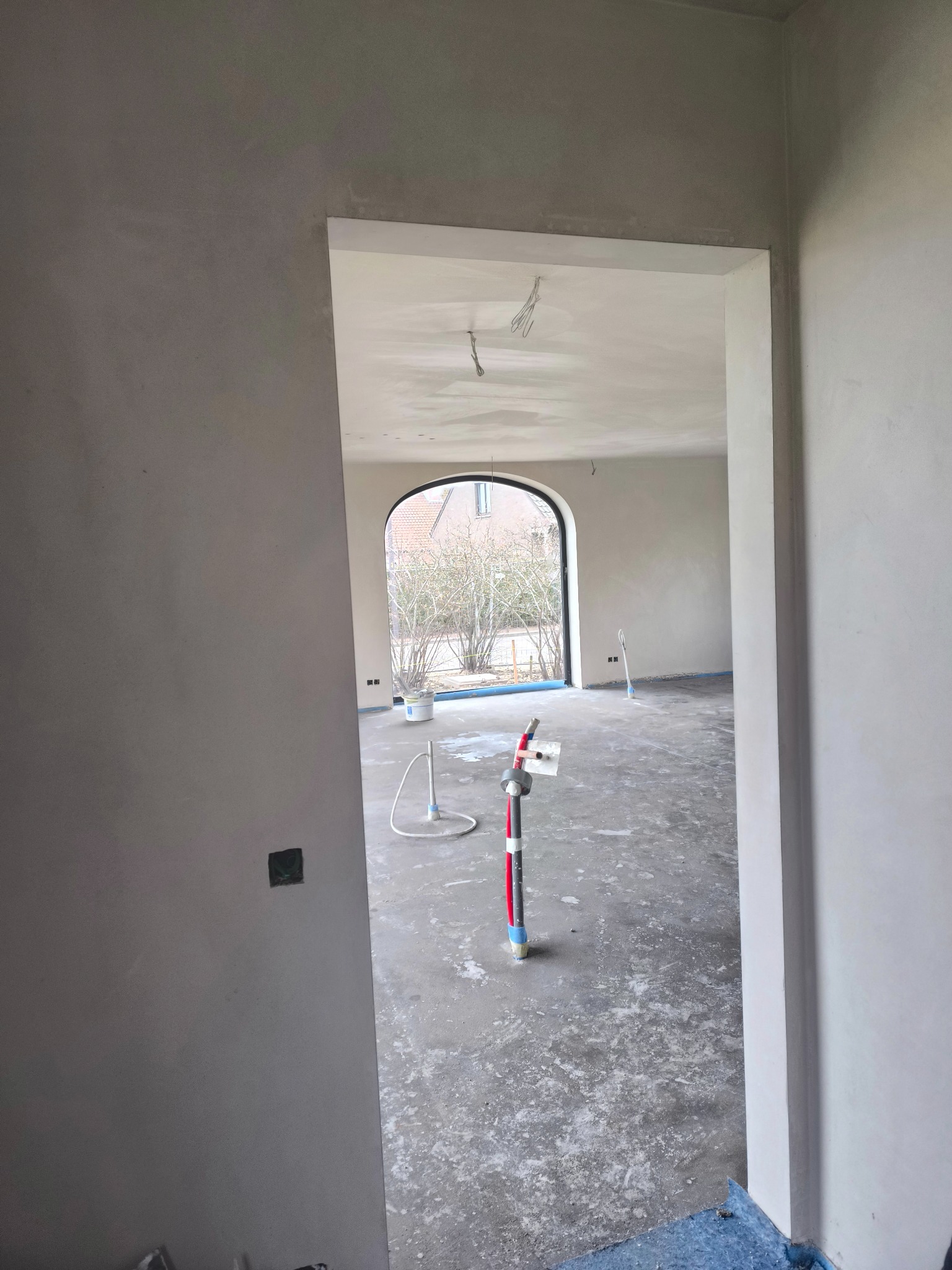 Wnętrze po szpachlowaniu, widok przez futrynę na jasne pomieszczenie z dużym oknem łukowym. Na podłodze widoczne narzędzia i resztki materiałów budowlanych.