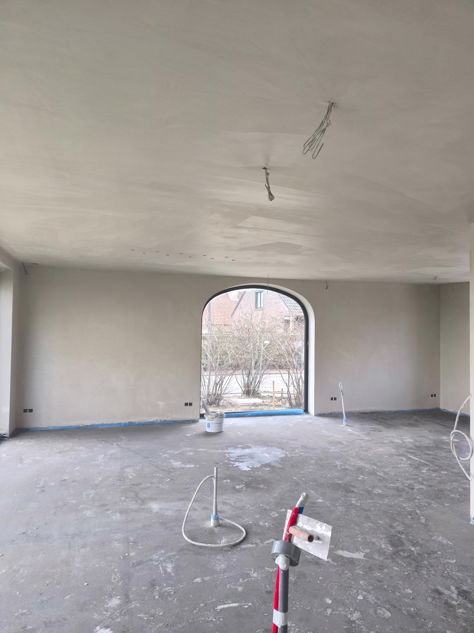 Wnętrze po szpachlowaniu: gładkie ściany i sufit w jasnym odcieniu, widoczne narzędzia pracy i zabezpieczenia podłogi. Duże okno łukowe z widokiem na zewnątrz.