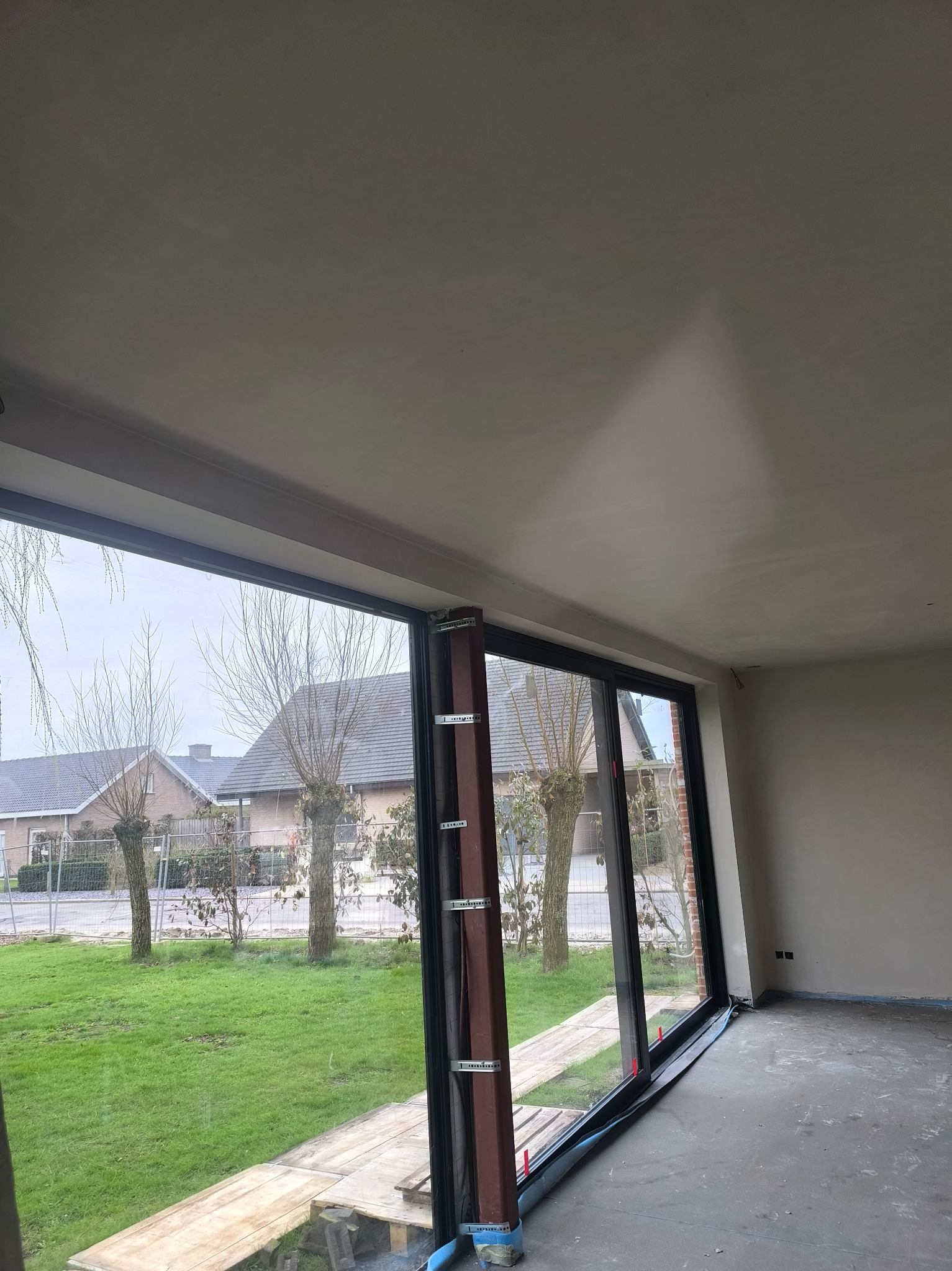 Surowe wnętrze z dużym oknem i świeżo wykończonymi ścianami, gotowe do dalszych prac wykończeniowych. Widok na zewnątrz z zielonym trawnikiem.