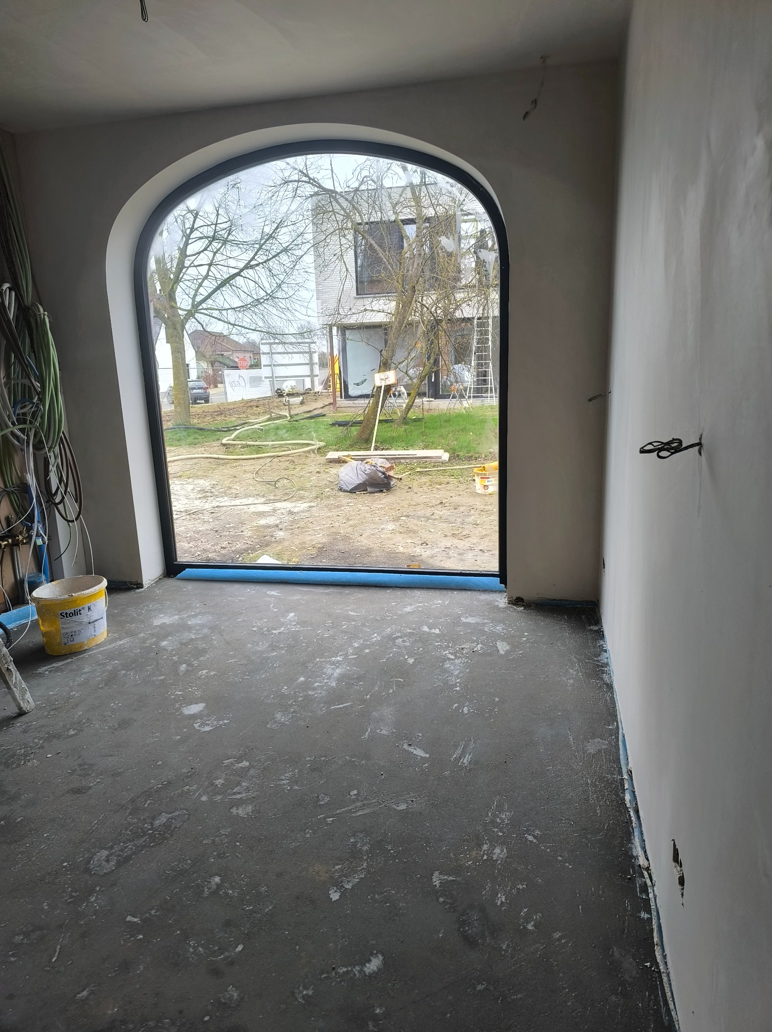 Wnętrze w trakcie prac wykończeniowych: surowe ściany, widoczne przewody elektryczne, okno łukowe z widokiem na zewnątrz. Wiadro z farbą w rogu pomieszczenia.