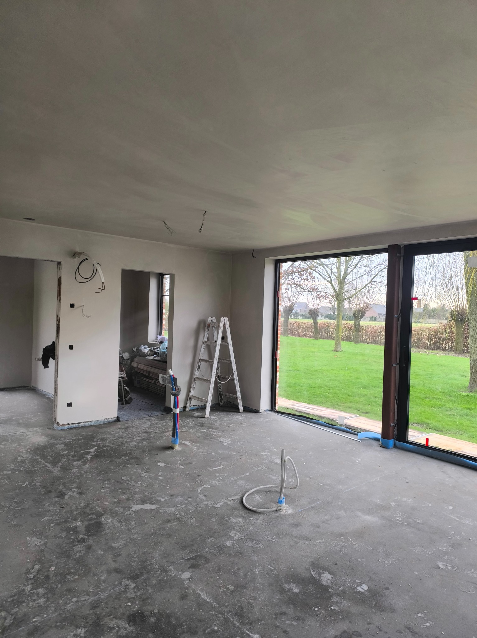 Surowe wnętrze w trakcie prac wykończeniowych: gładkie ściany, widoczne instalacje, drabina oparta o ścianę, duże okno z widokiem na zieleń. Podłoga betonowa.