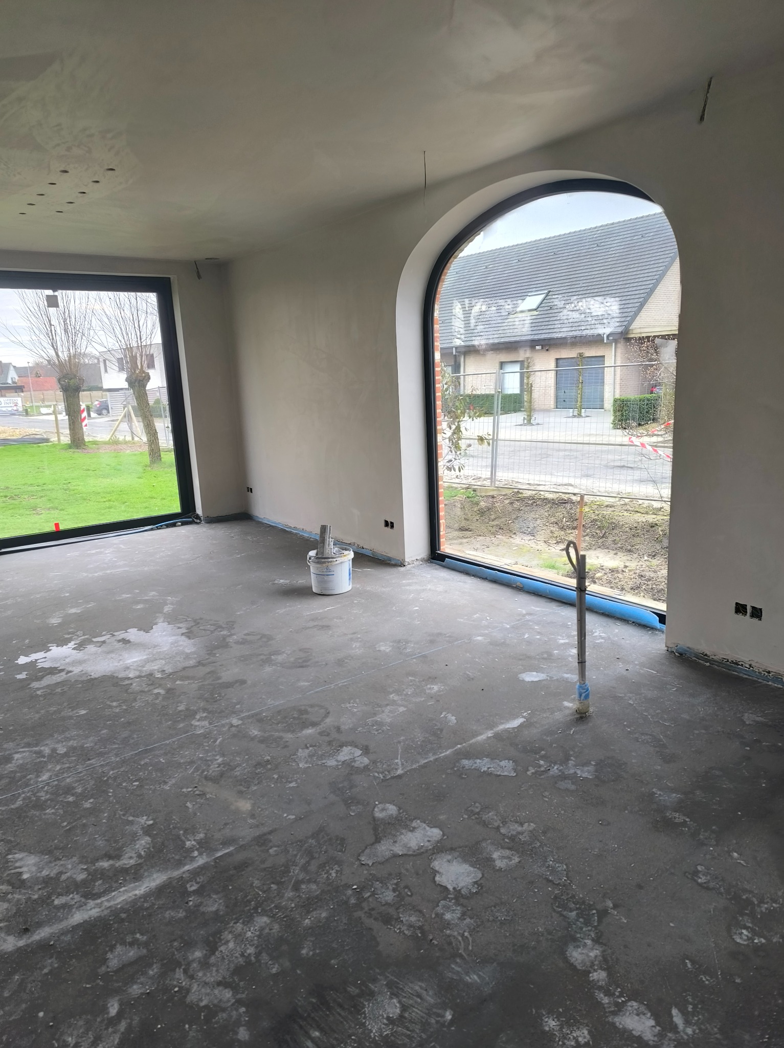 Wnętrze w trakcie prac wykończeniowych: ściany po szpachlowaniu, przygotowane do dalszych prac. Widoczne narzędzia i wiadro z materiałem. Duże okna z widokiem na zewnątrz.