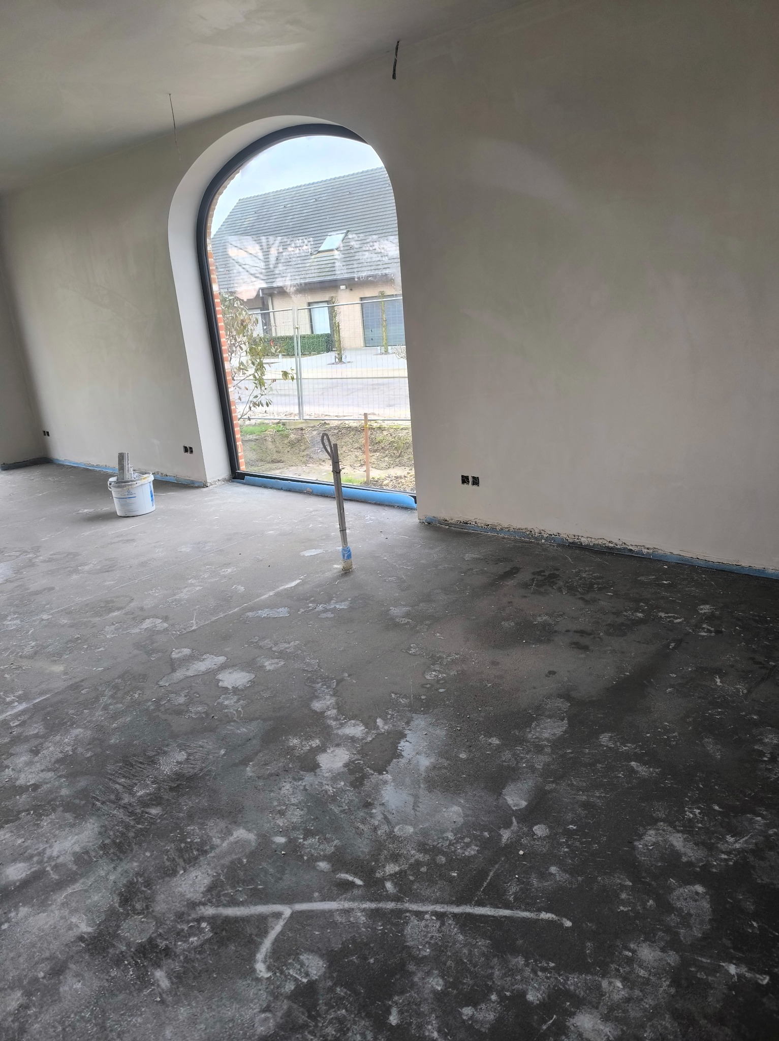 Wnętrze w trakcie prac wykończeniowych: ściany po nałożeniu gładzi, zabezpieczona podłoga, okno łukowe z widokiem na zewnątrz, wiadro z zaprawą i narzędzie.
