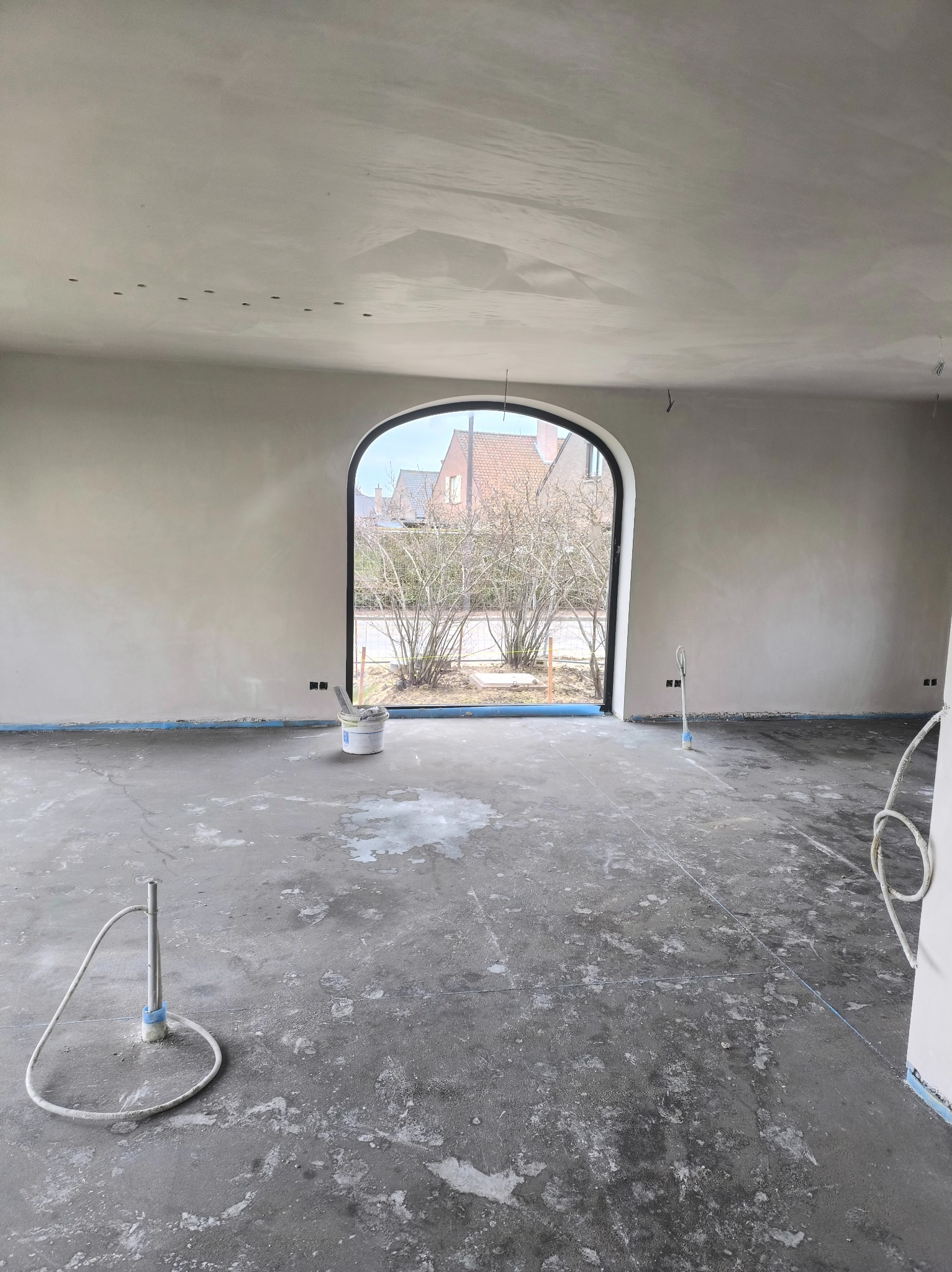 Wnętrze po szpachlowaniu ścian i sufitu, widoczne ślady po pracach na podłodze, okno łukowe z widokiem na zewnątrz, przygotowane do dalszych prac wykończeniowych.