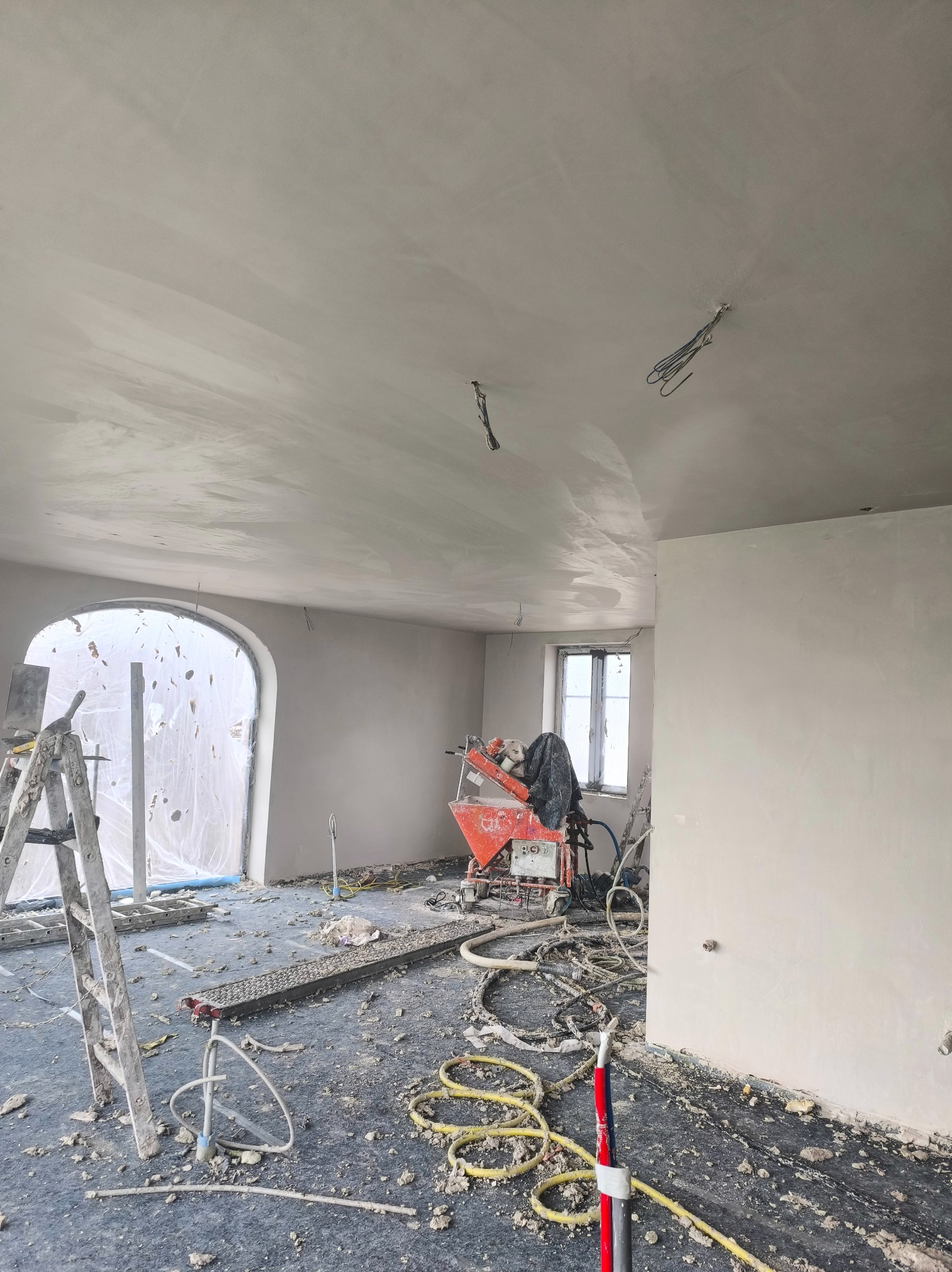 Wnętrze w trakcie prac wykończeniowych: ściany i sufit pokryte świeżą warstwą gładzi, widoczna maszyna do nakładania gładzi, drabina i zabezpieczone okno.