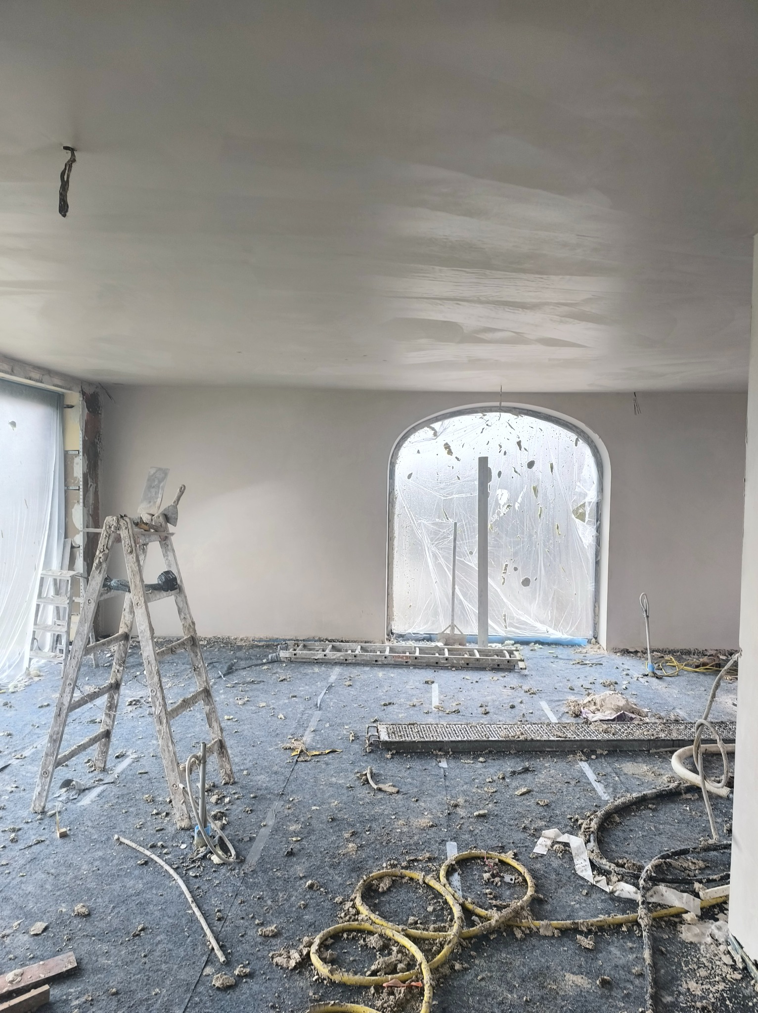 Wnętrze w trakcie prac wykończeniowych: ściany po nałożeniu gładzi, zabezpieczone okno łukowe, drabina malarska i akcesoria na podłodze.