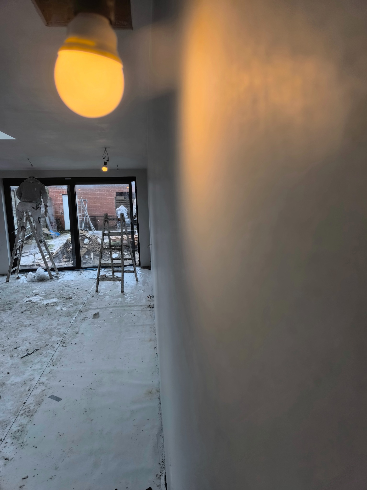 Wnętrze w trakcie prac wykończeniowych: ściana po szpachlowaniu, zabezpieczona podłoga, drabiny i pracownik w kombinezonie malarskim w tle, oświetlone żarówką.