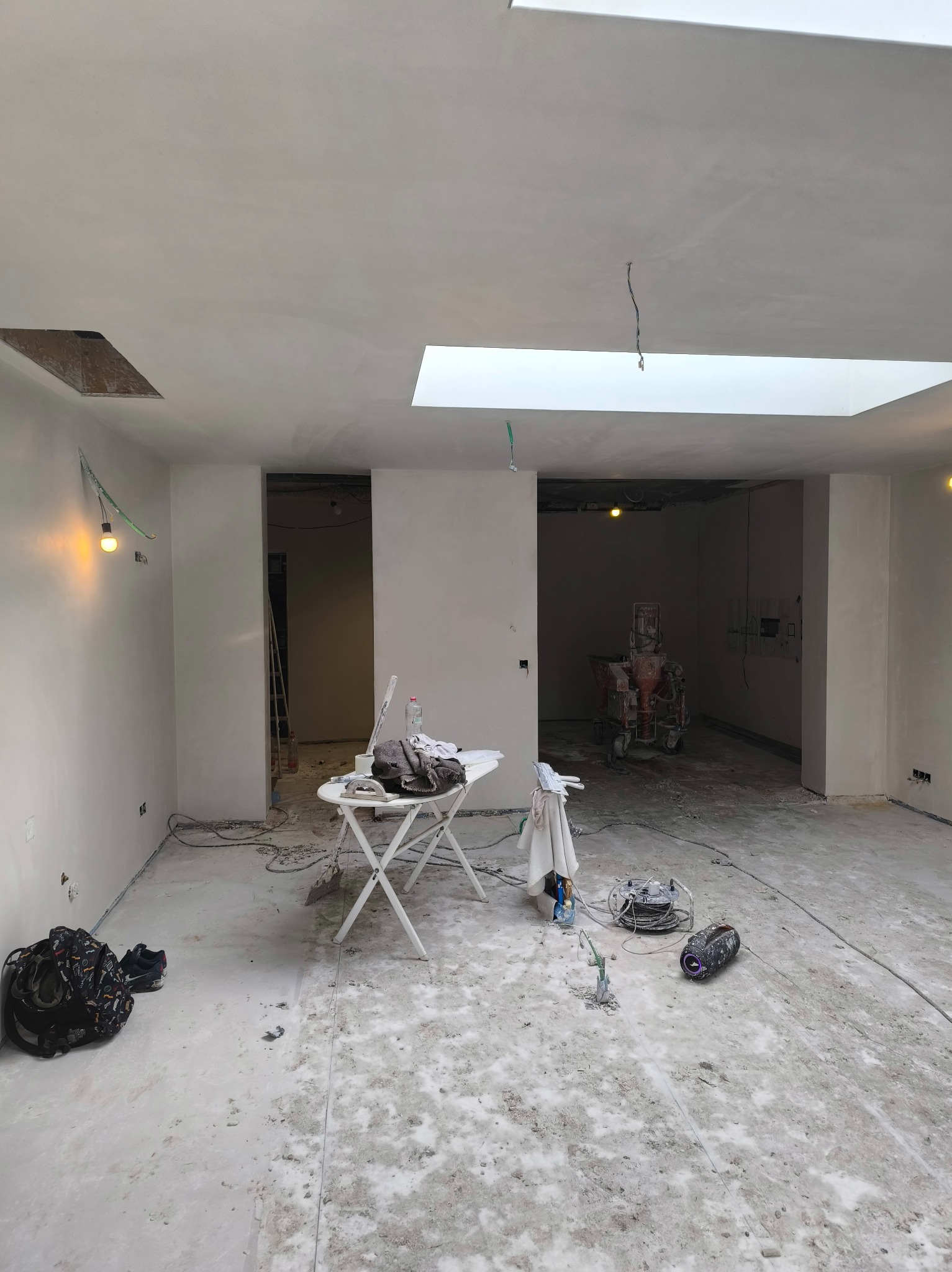 Wnętrze po szpachlowaniu: białe ściany, sufit z oknami dachowymi, sprzęt budowlany, stół roboczy. Widoczne ślady prac na podłodze. W tle maszyna do tynkowania.