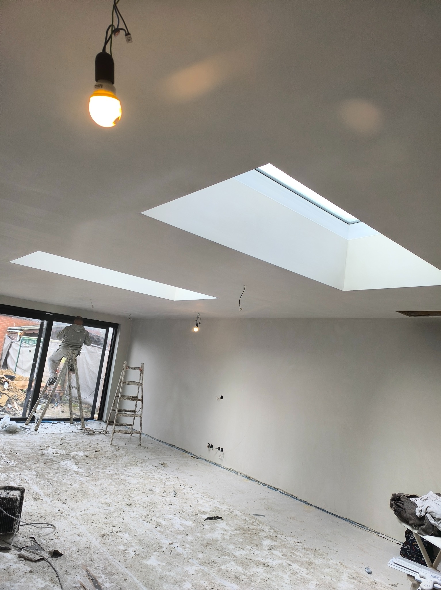 Praca w toku: malarz na drabinie maluje jasne wnętrze z oknami dachowymi. Podłoga zabezpieczona folią, widoczne ślady farby. Żarówka zwisa z sufitu.