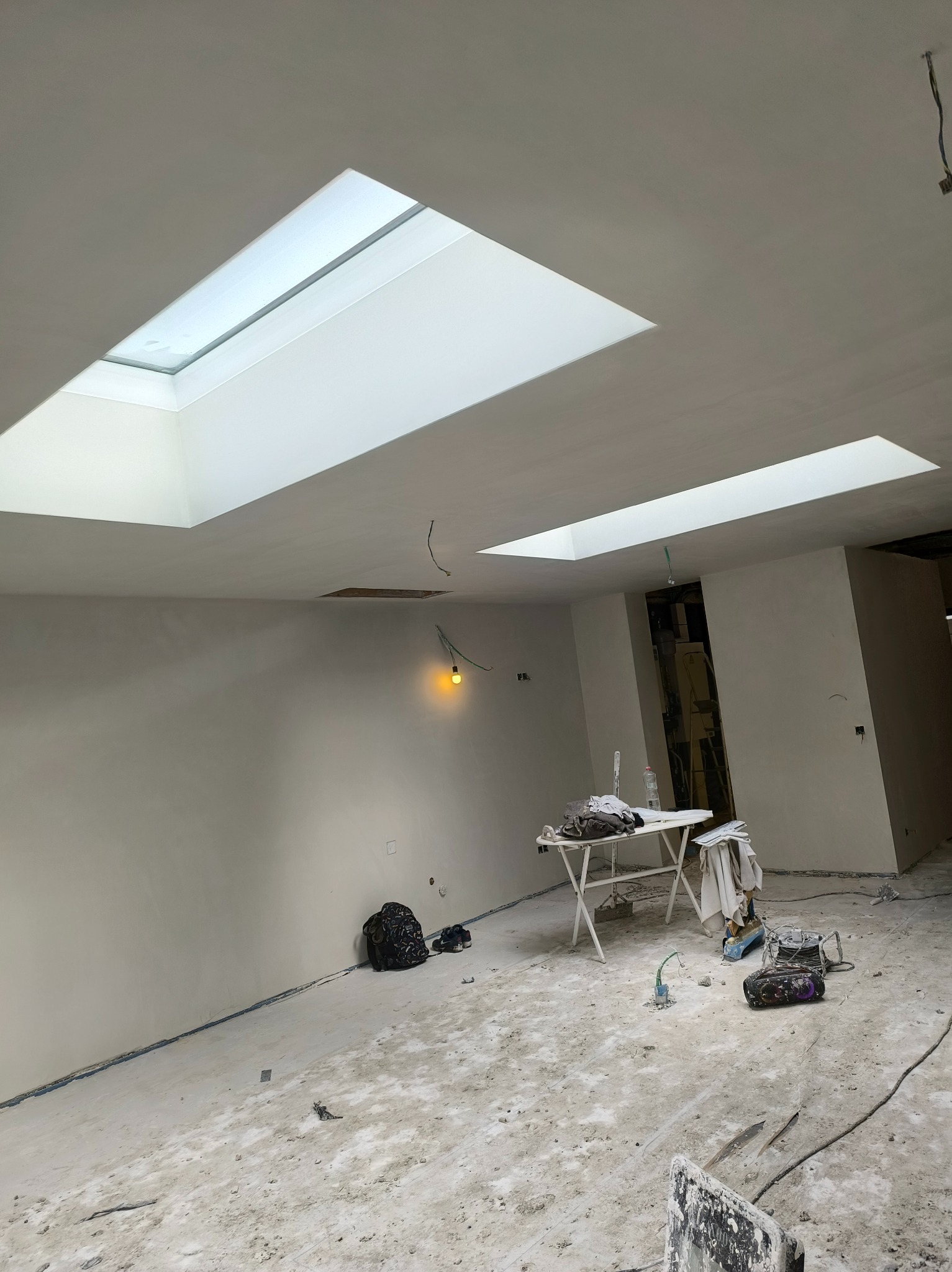 Jasne wnętrze po pracach wykończeniowych z oknami dachowymi. Widoczne narzędzia, stół roboczy i ślady po szpachlowaniu na podłodze. Ściany przygotowane do malowania.