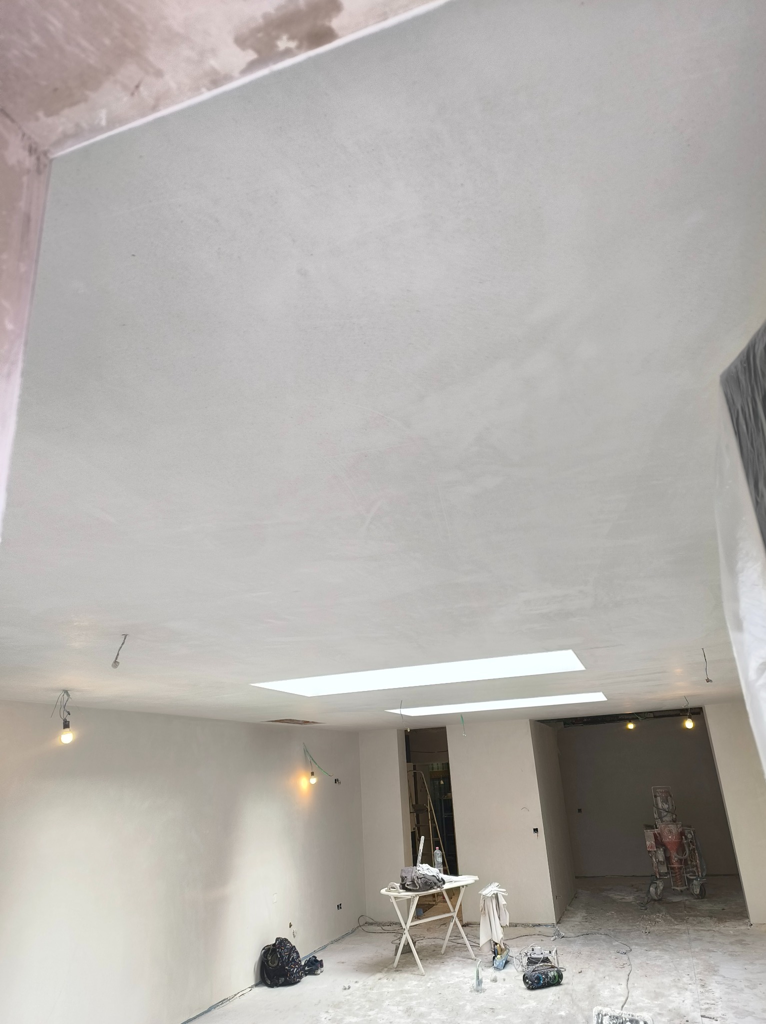 Wnętrze po nałożeniu gładzi: jasne ściany i sufit z oknami dachowymi, widoczne narzędzia i maszyna do nakładania gładzi. Minimalistyczny wystrój, surowe wykończenie.