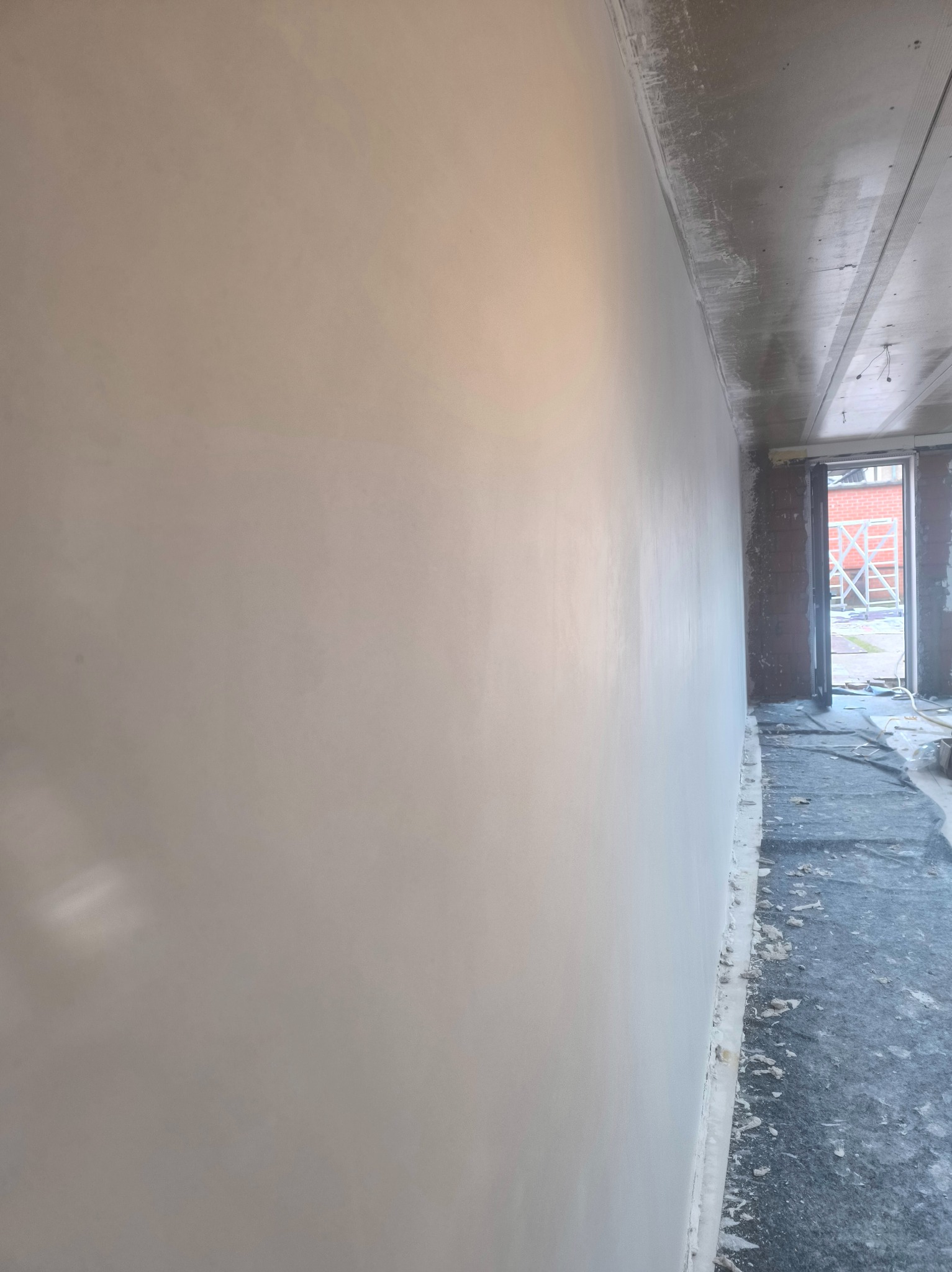 Gładka, jasna ściana po szpachlowaniu w surowym wnętrzu. Widoczne zabezpieczenie podłogi i fragment rusztowania w tle. Świeżo wykończona powierzchnia.